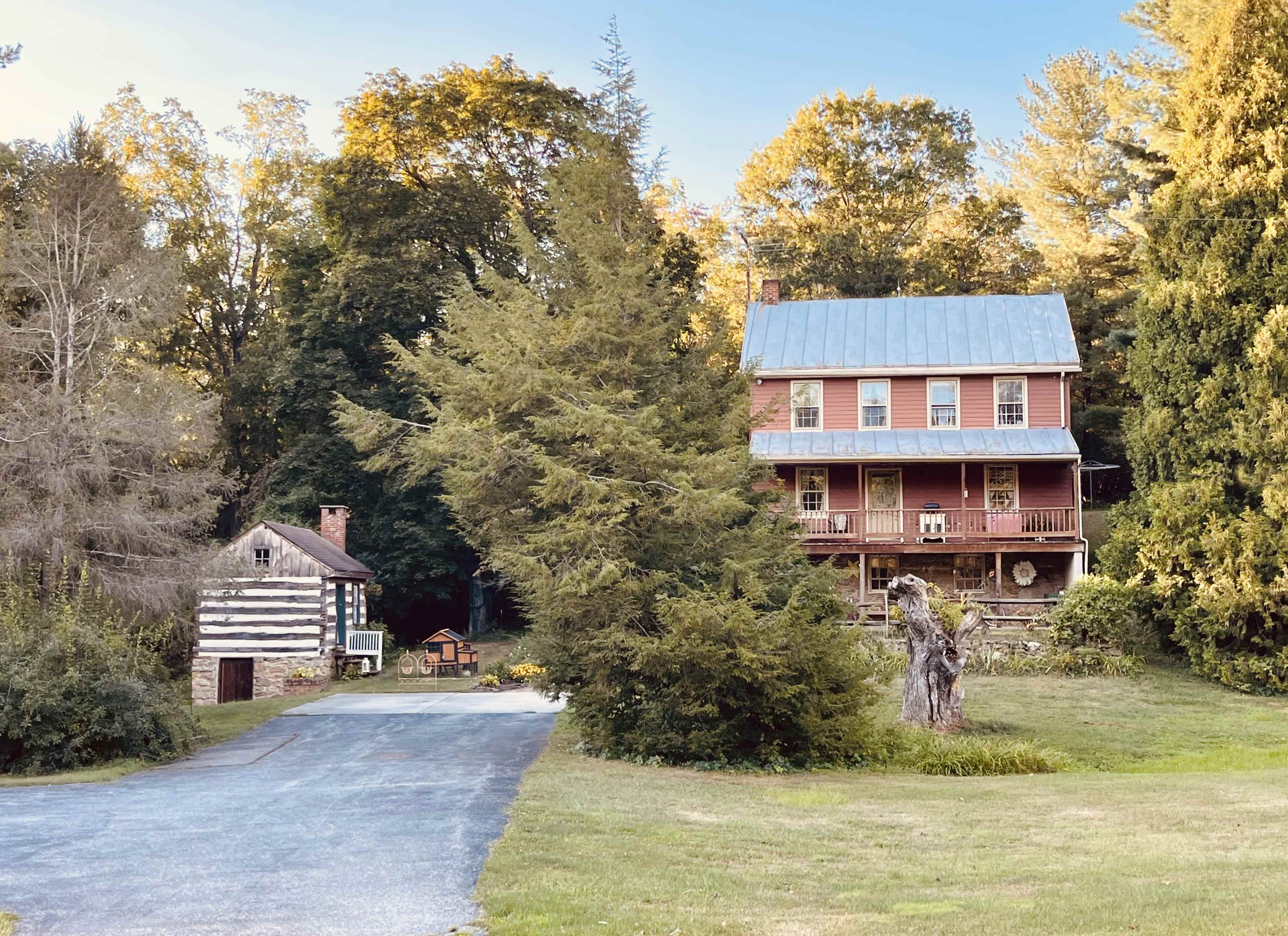 The image shows a rustic house with a blue metal roof and a log cabin beside it, set in a wooded area with green grass and trees.