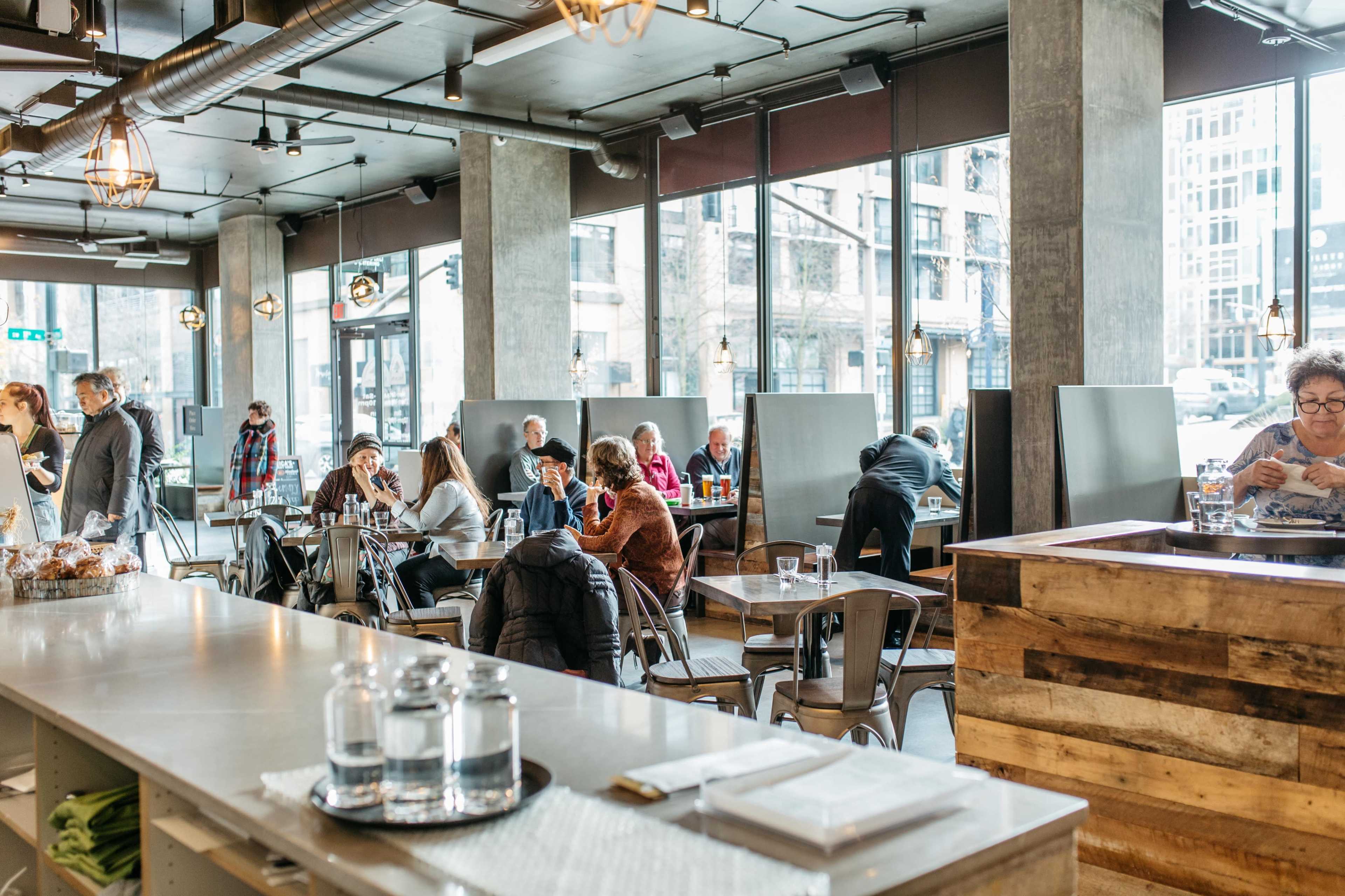 The image shows a busy modern café filled with patrons seated at various tables and a counter with water glasses in the foreground.