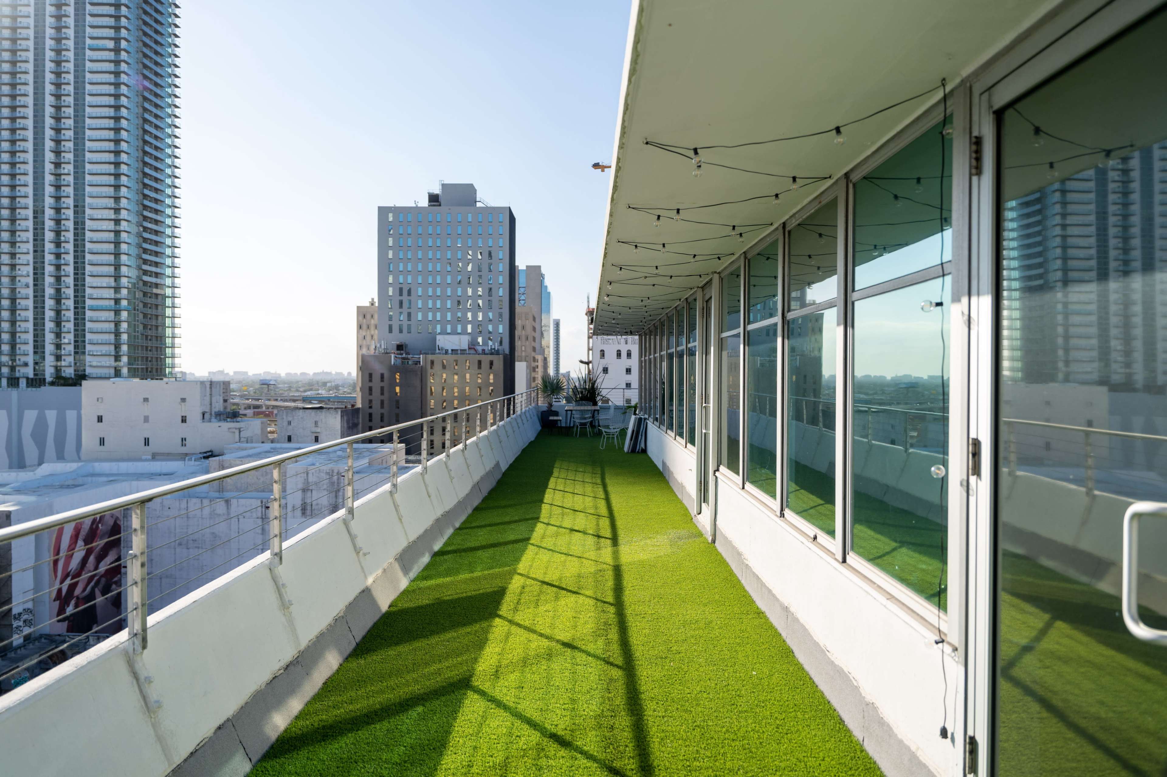 A long, green, artificial grass walkway runs alongside a modern building, with city skyscrapers visible in the background under clear skies.