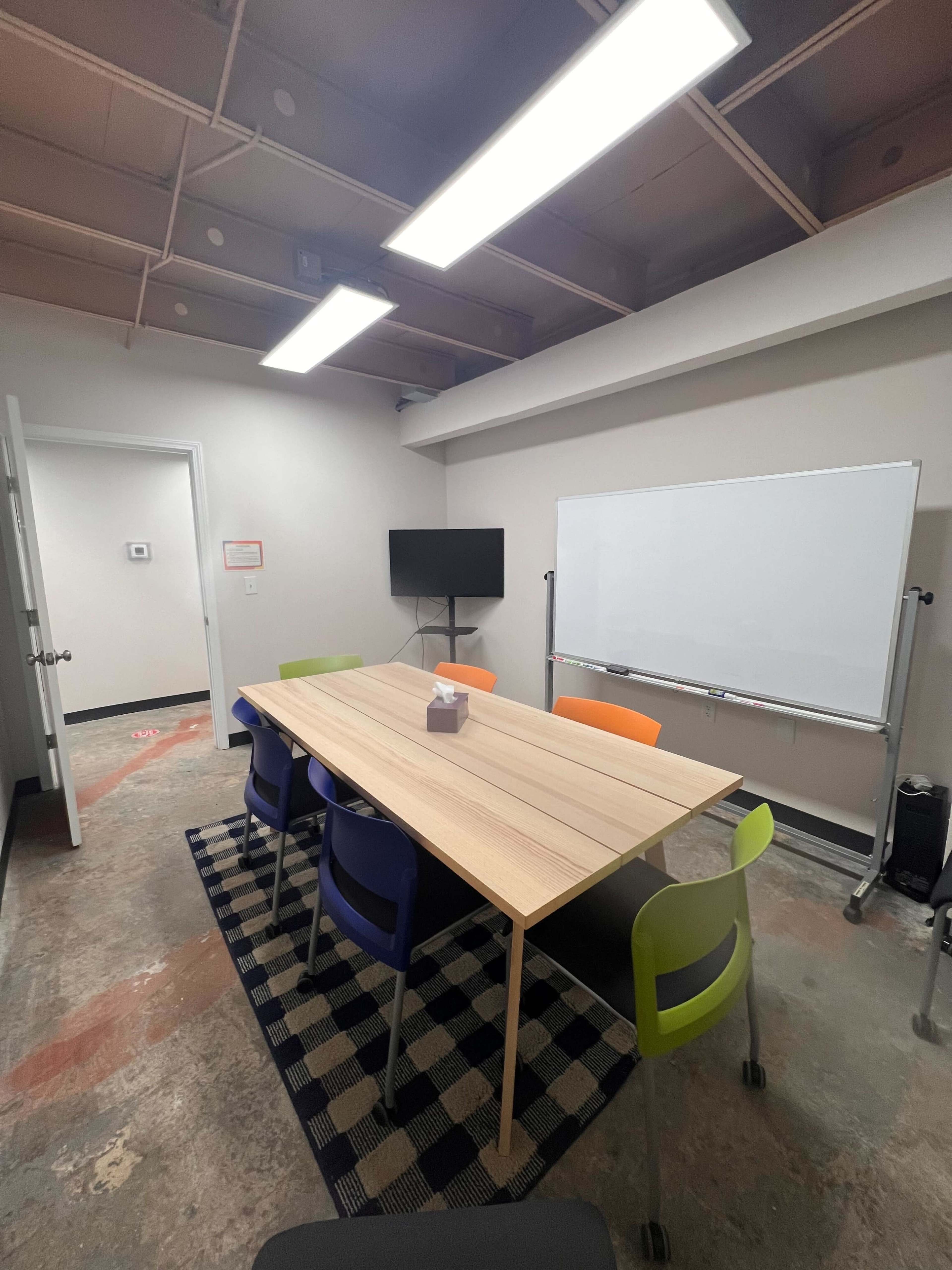 A rectangular table with six colorful chairs is set in a meeting room that features a whiteboard and a television on the wall.