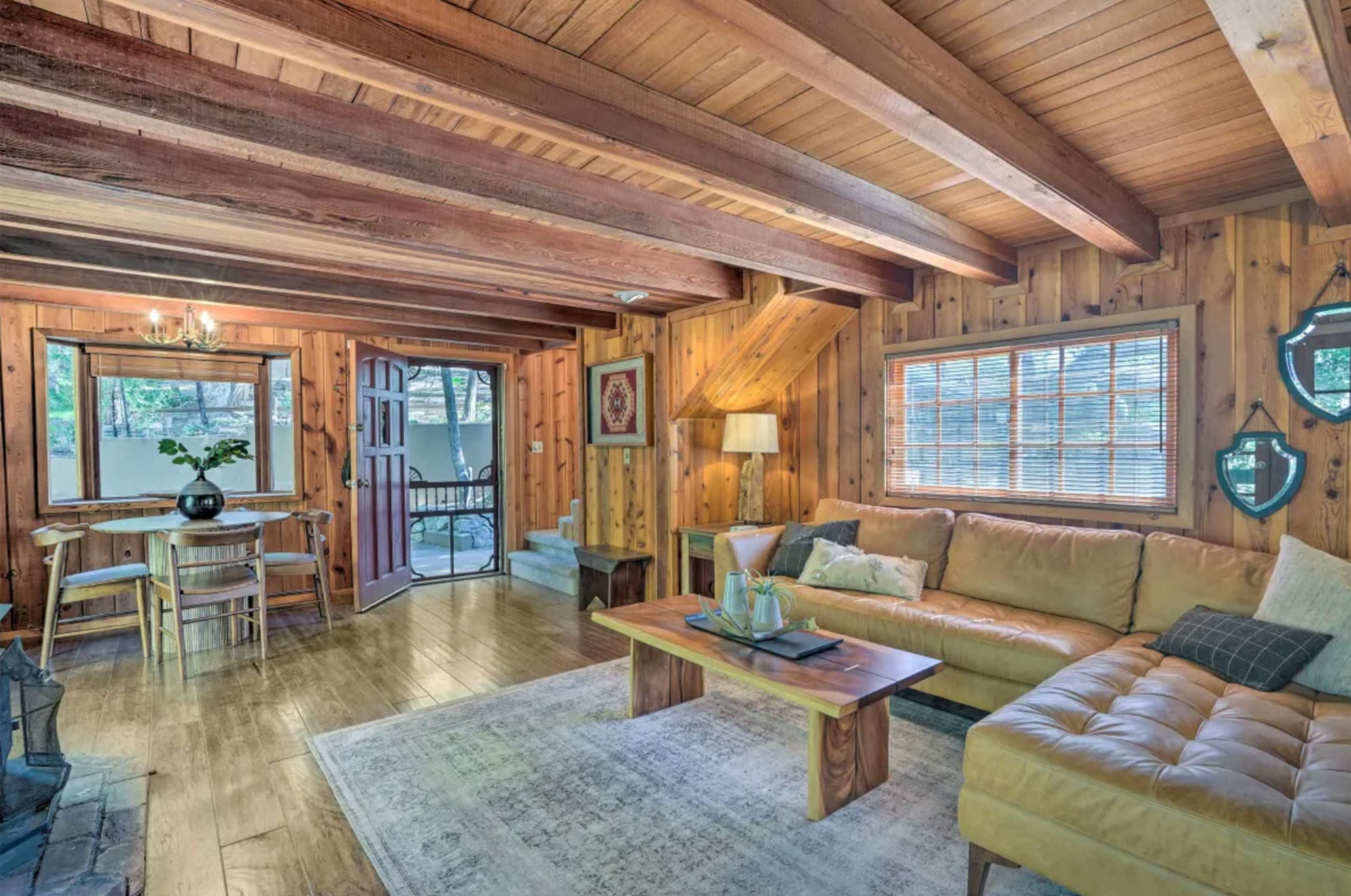 The room features wooden beams and walls, a tan sofa, a coffee table, a dining area, and a door leading outside.