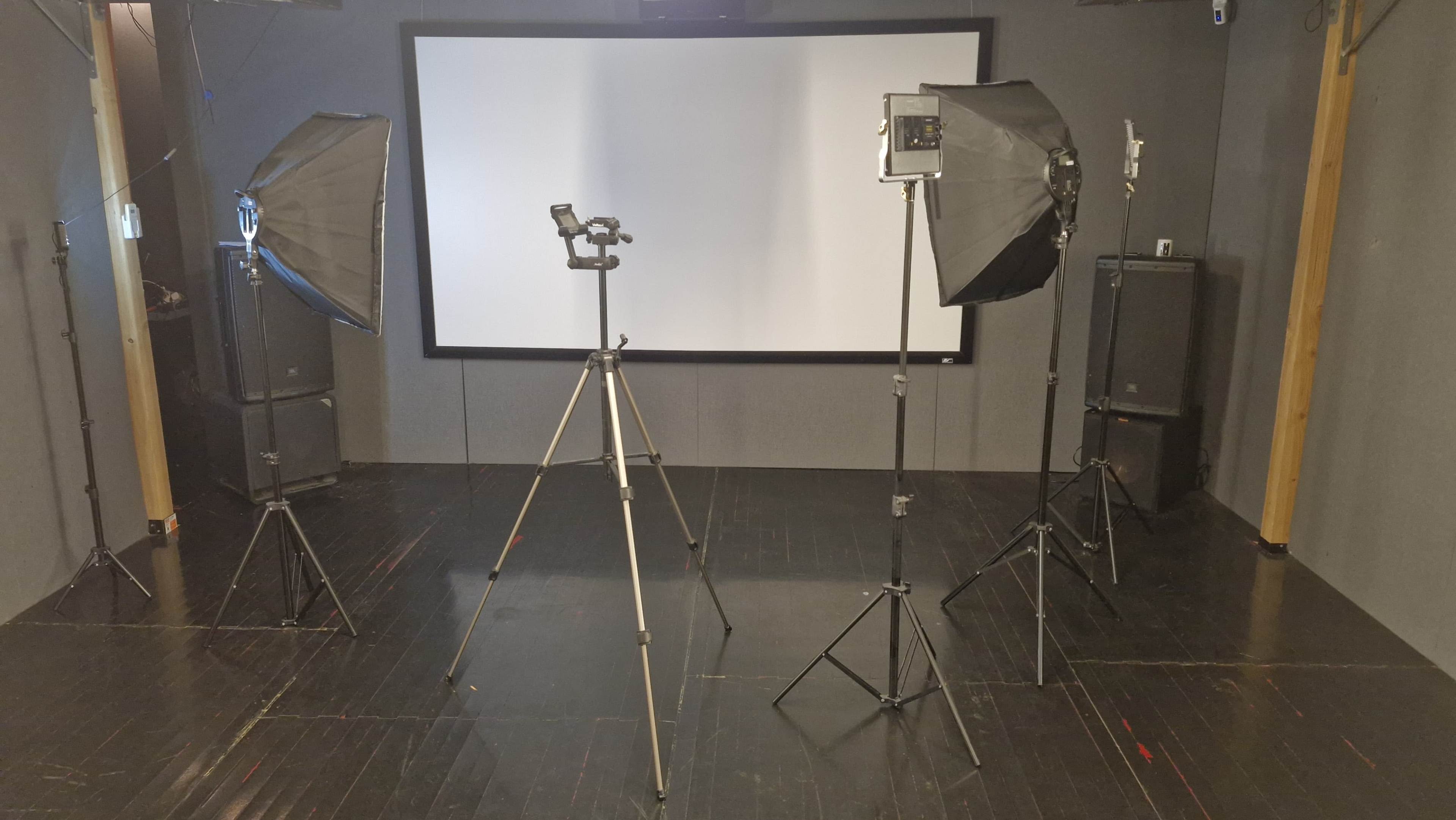 A photo studio setup featuring two softbox lights, a camera on a tripod, and a large projection screen in a dark room.