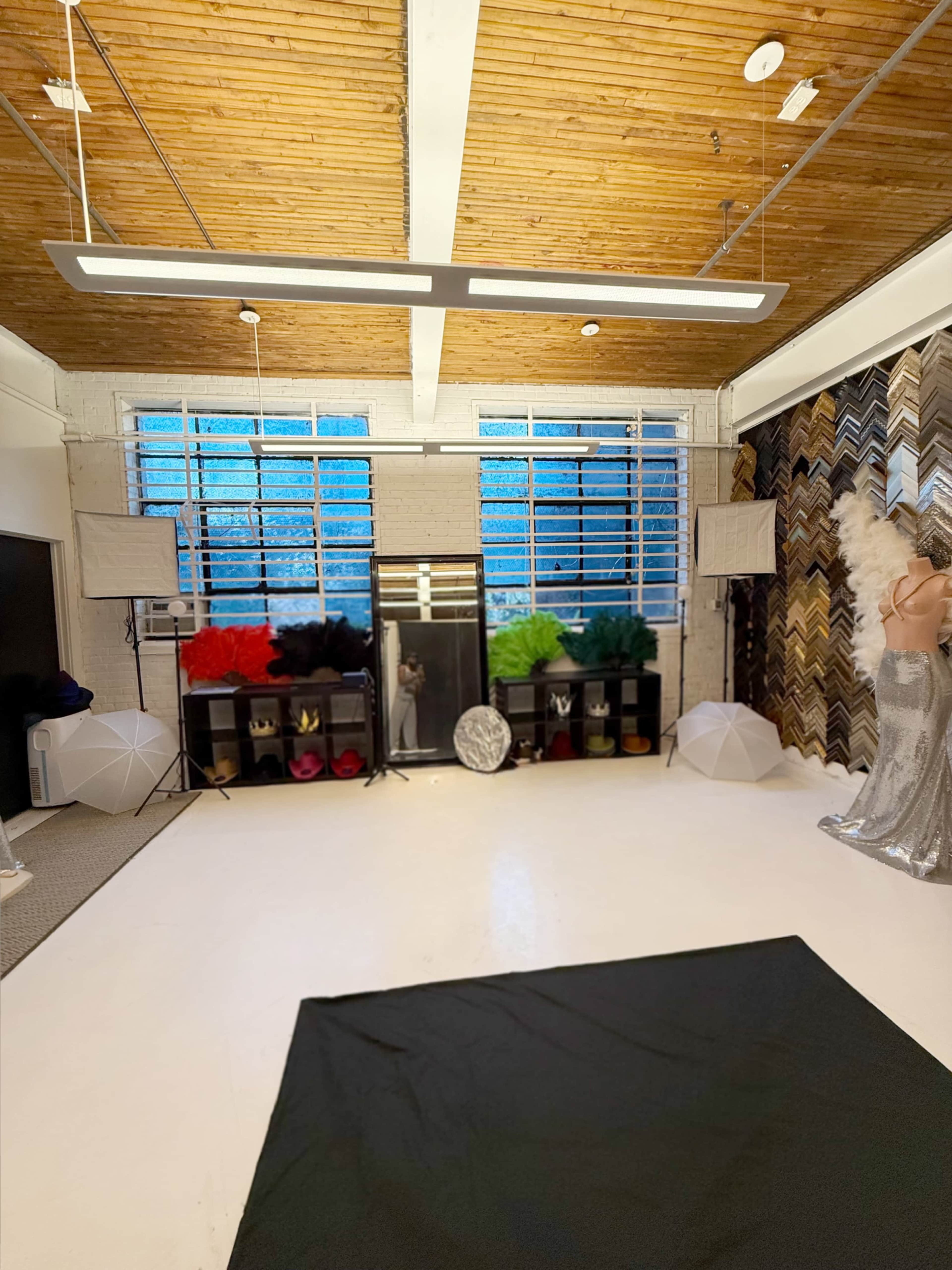 The image shows a spacious indoor studio with a white floor, large windows, various props on shelves, and a mannequin wearing a silver outfit.