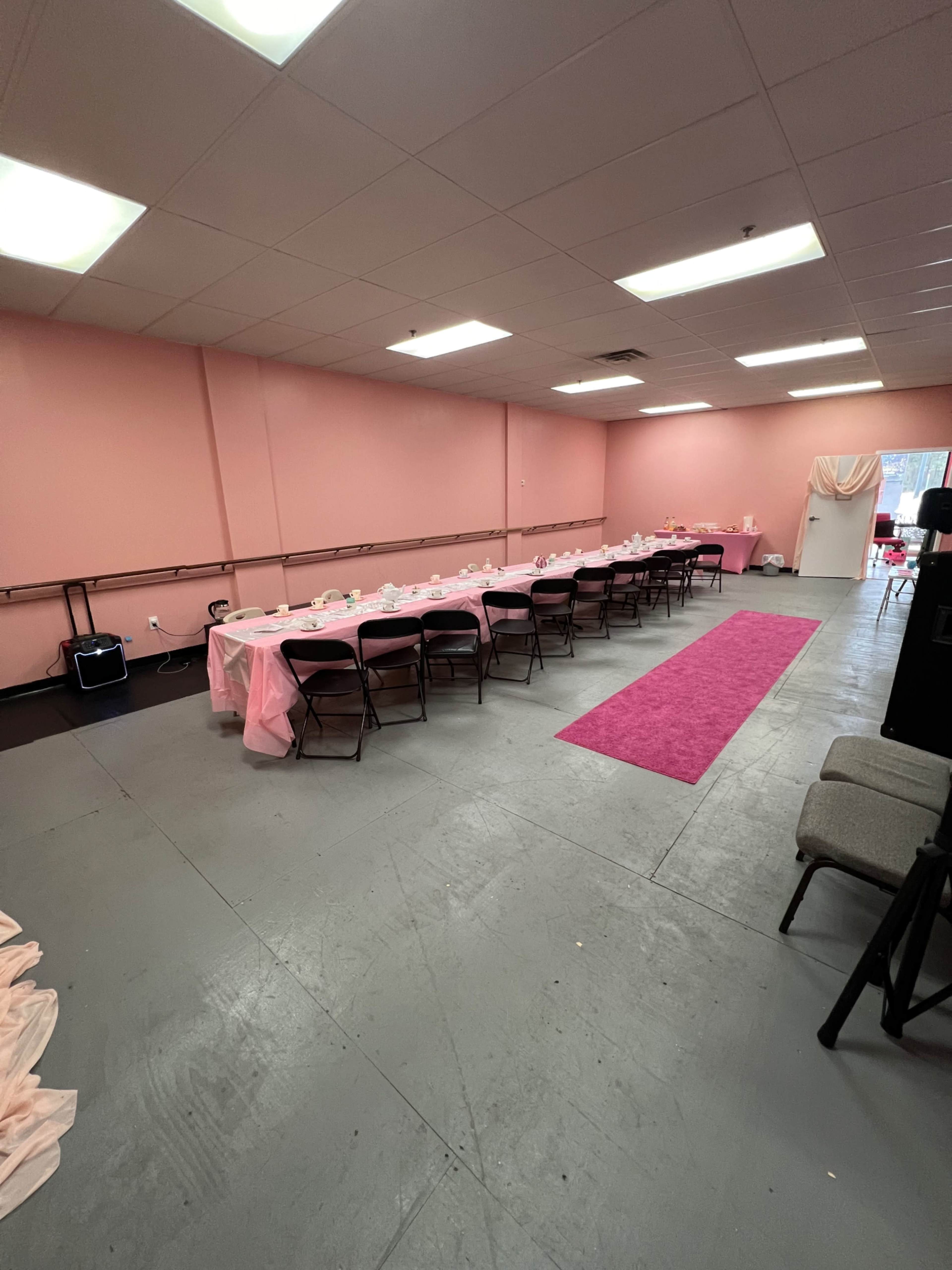 A pink-themed event space features a long table set with chairs, centered on a pink carpet, against a backdrop of pink walls.