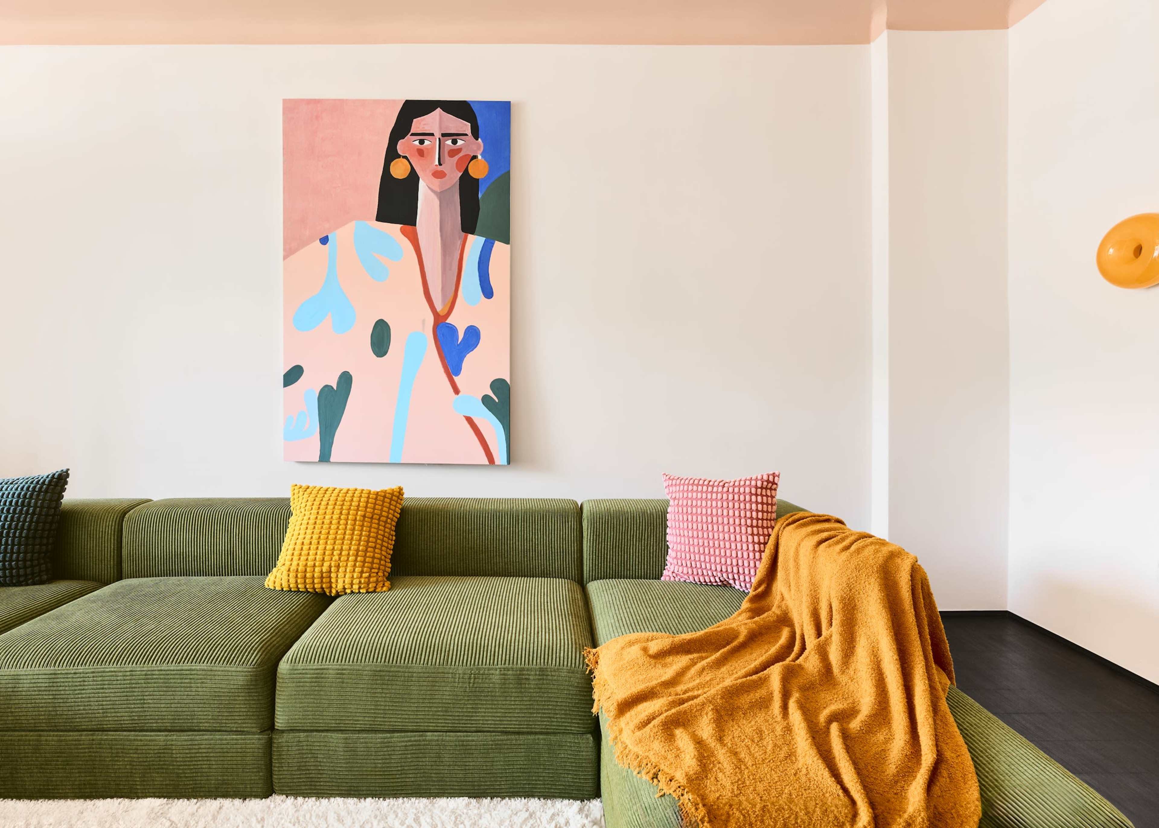 The image shows a modern living room with a green sectional sofa, colorful throw pillows, a cozy orange blanket, and a large abstract portrait hanging on the white wall.