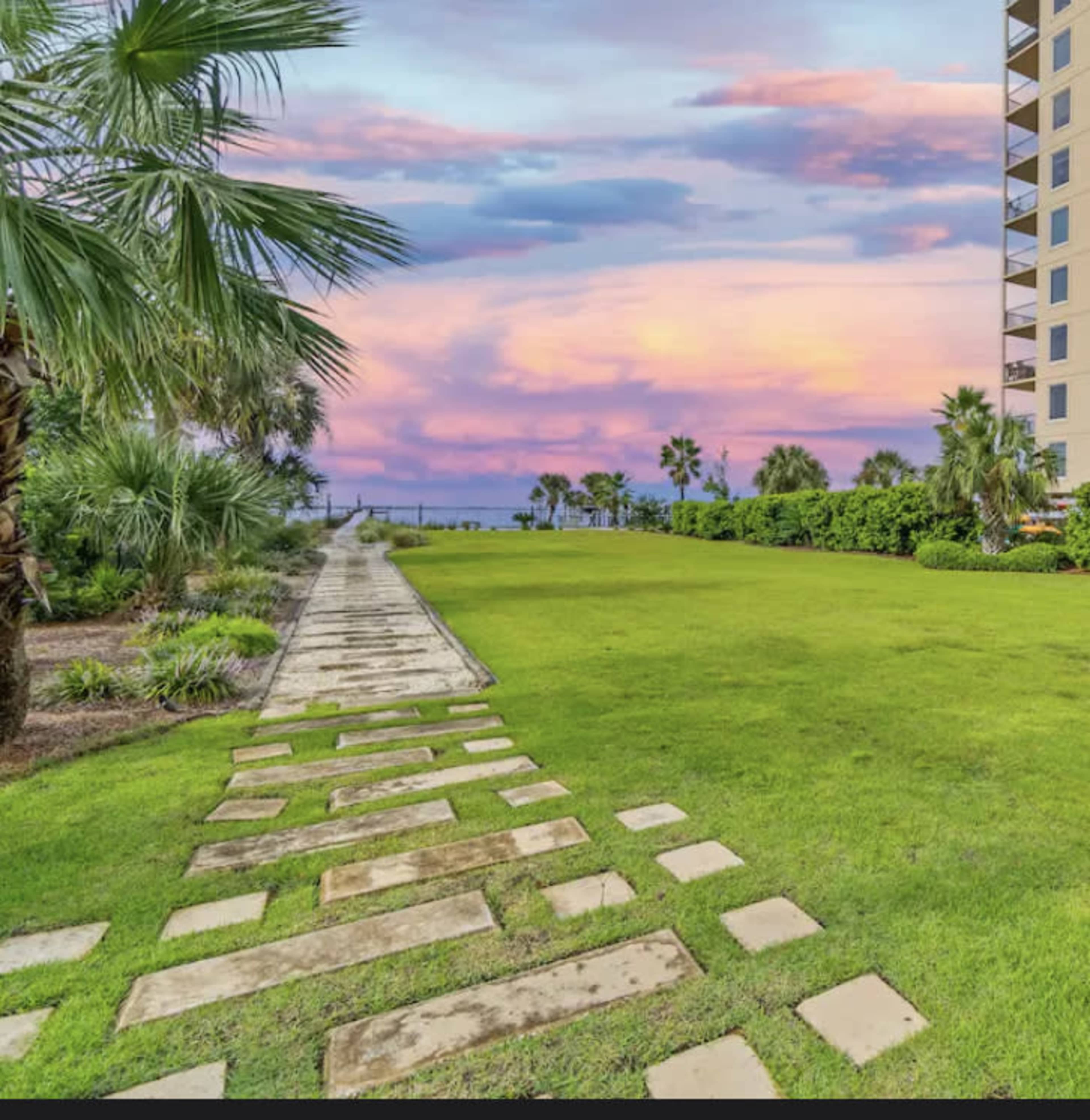 A stone pathway leads through a grassy area bordered by palm trees, with a colorful sunset reflected in the sky above the ocean in the background.