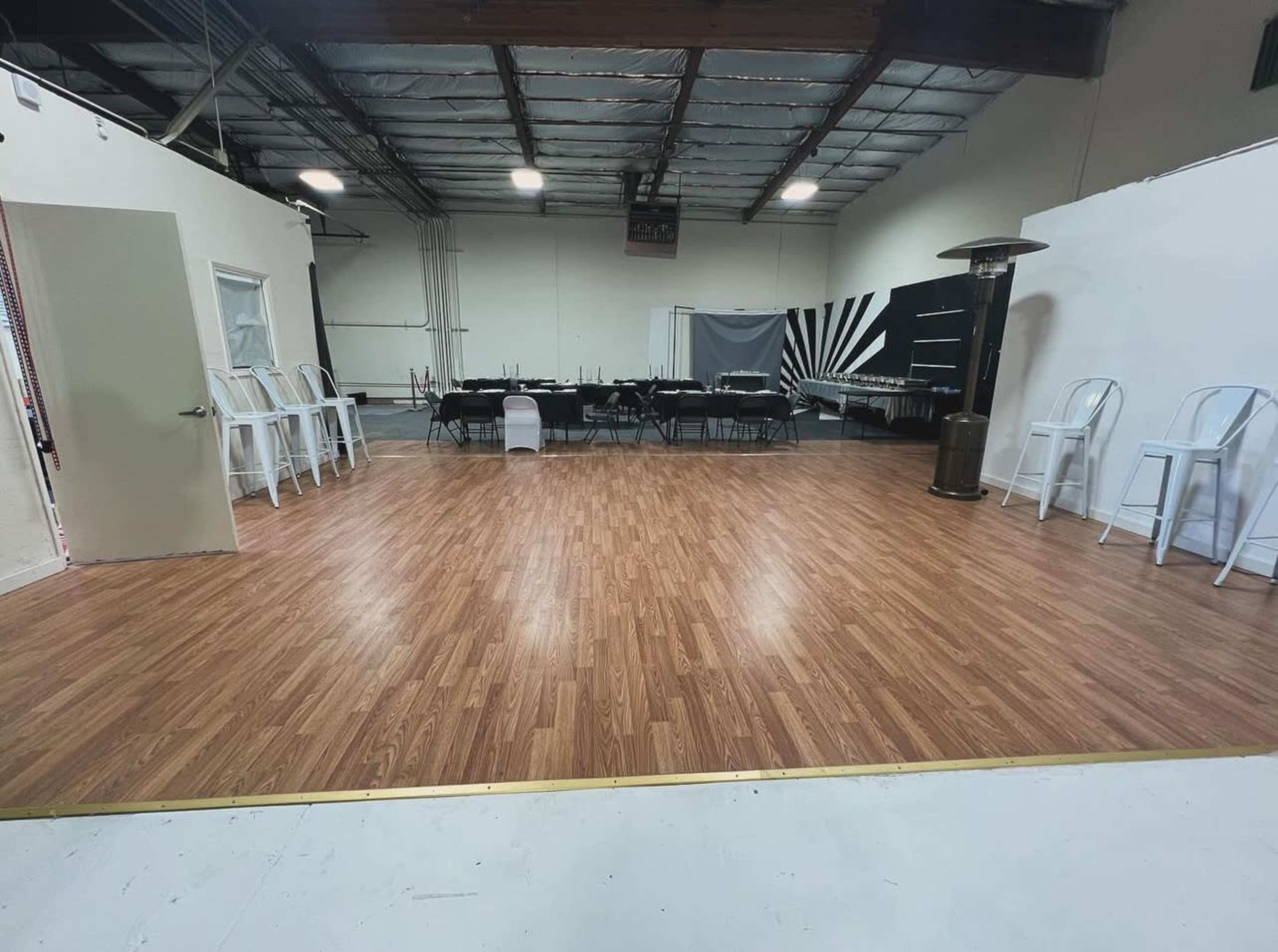 A spacious interior room with a polished wood floor, a long black table set up for seating, and a few chairs arranged along the walls.