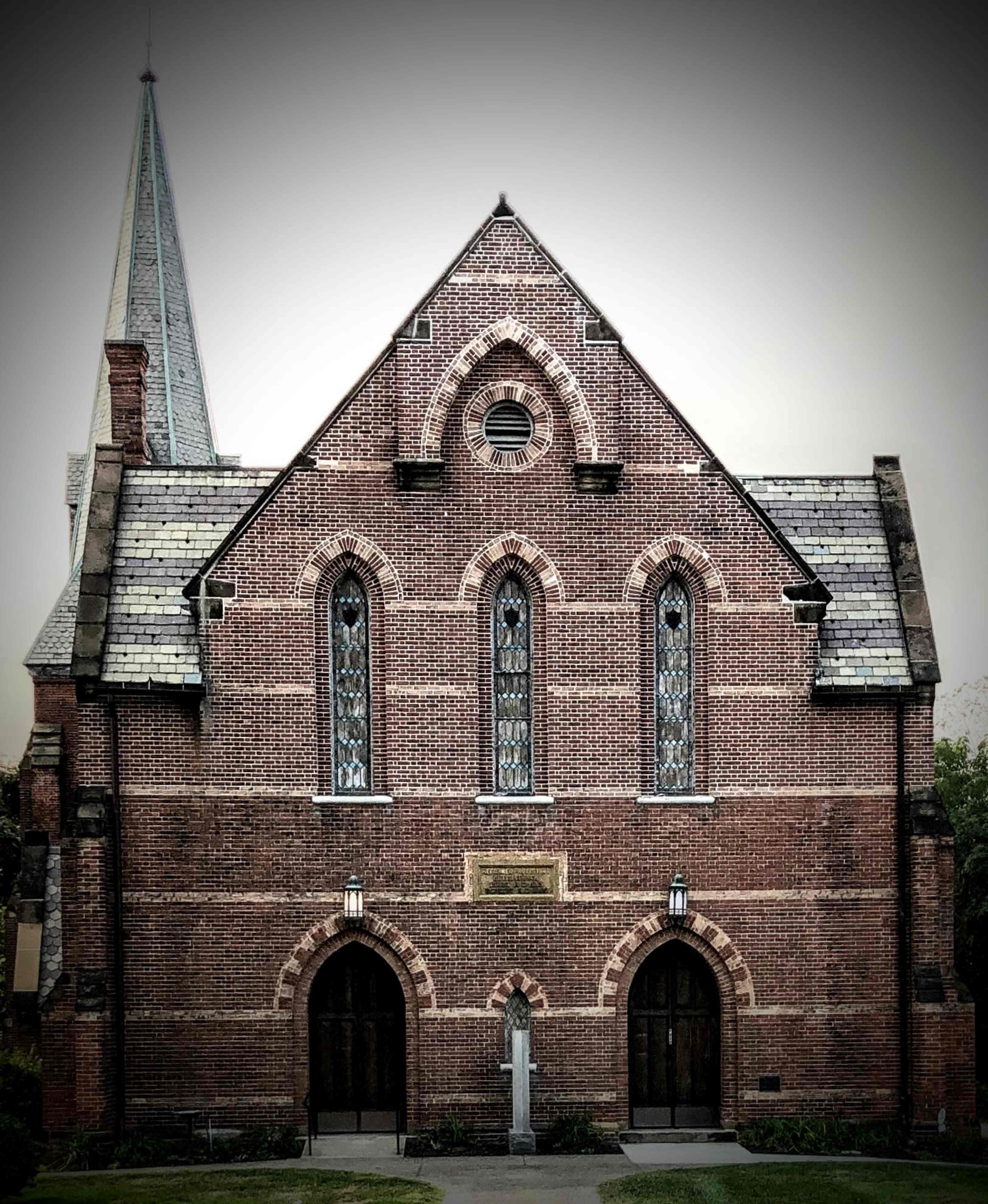 A brick church with a steep roof, pointed arch windows, and a spire, set against a muted sky.