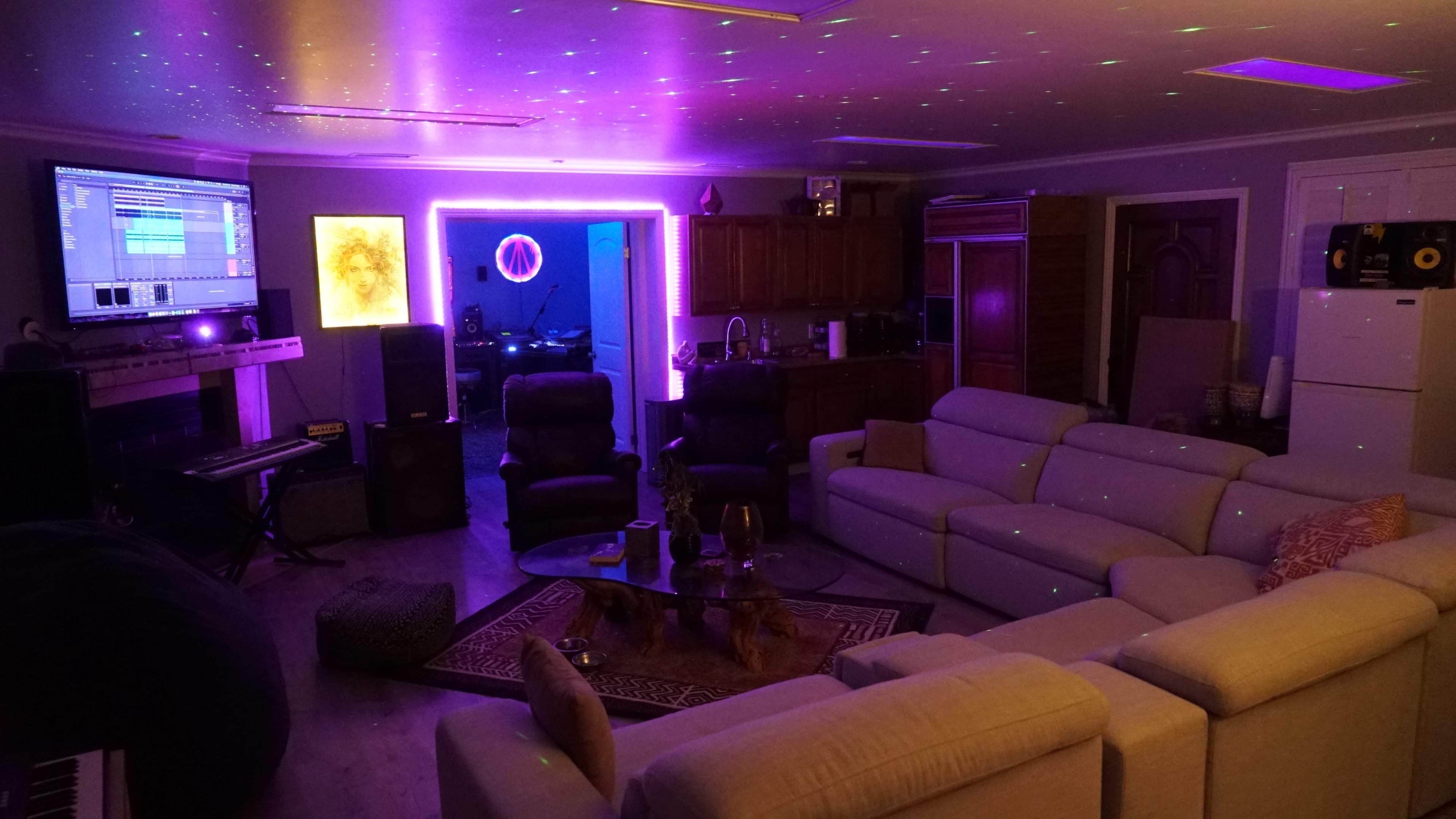 A living room featuring a large white sectional sofa, a coffee table, and a screen displaying music software, illuminated by multicolored lights and a purple glow.