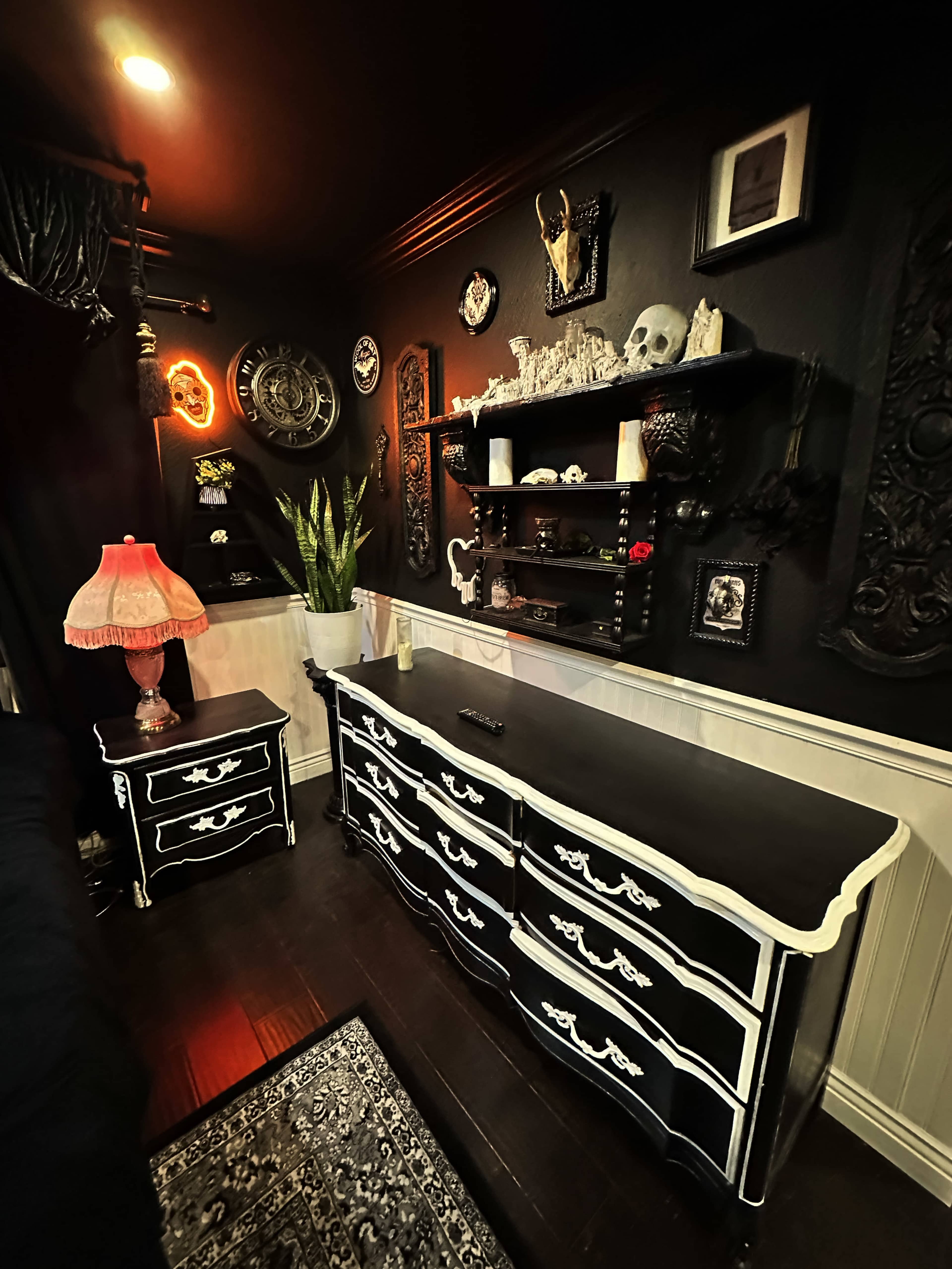 The image shows a dimly lit room featuring a black dresser with white accents, a matching nightstand, a decorative wall shelf, and various decorative items against a dark backdrop.