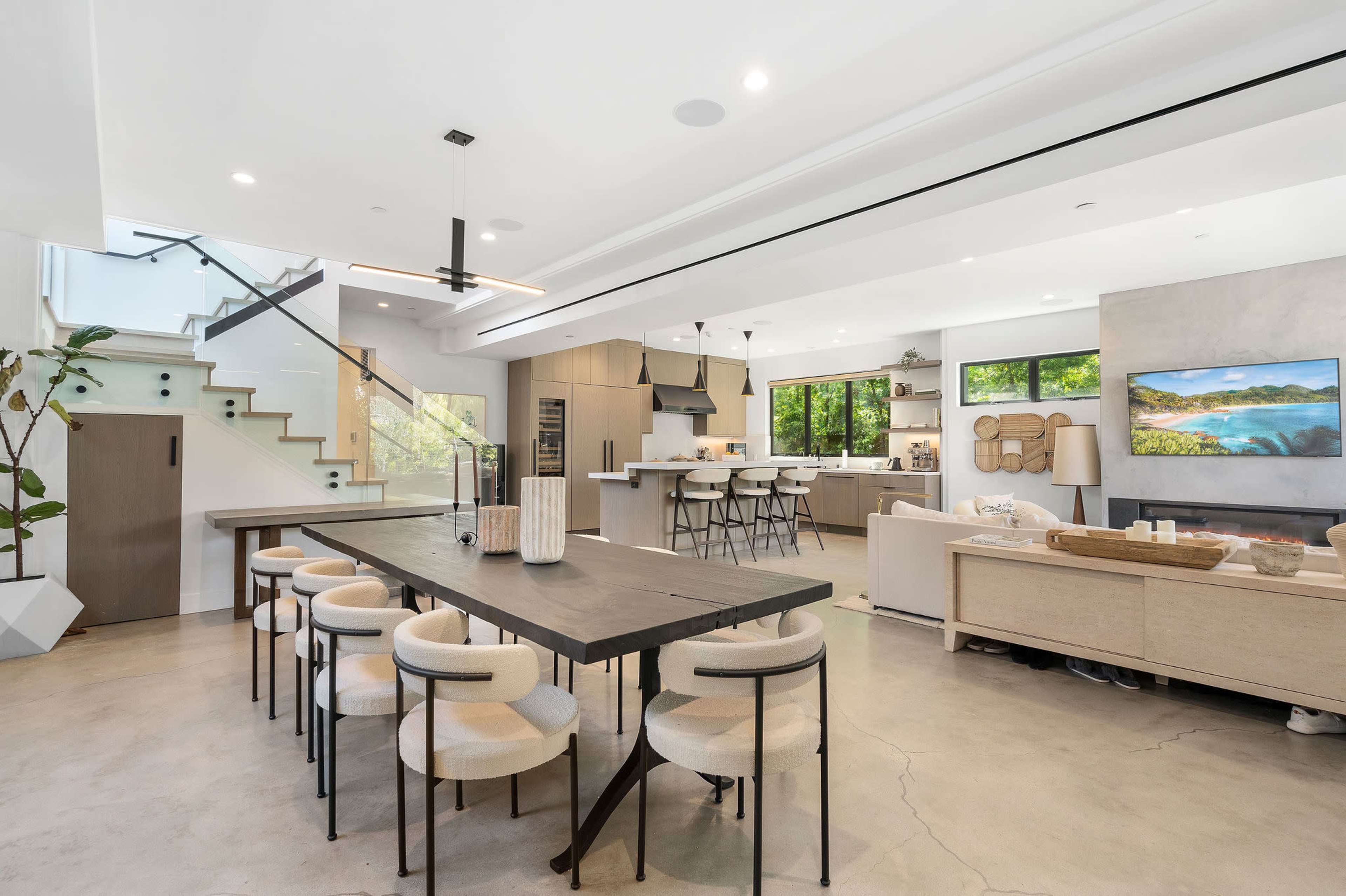 A modern open-concept living space with a dining area, kitchen, and living room, featuring a large table, bar stools, and ample natural light.
