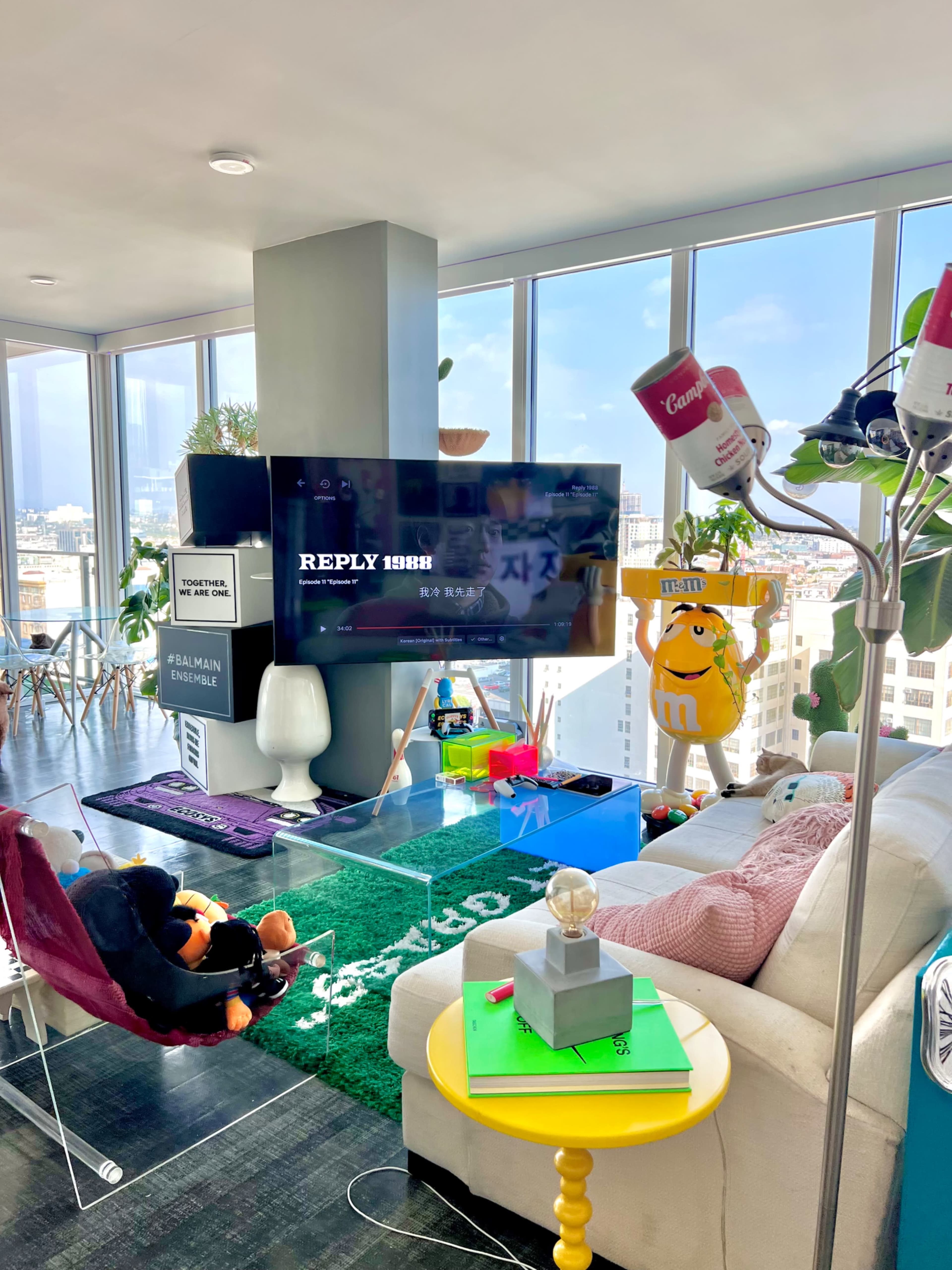 The image shows a modern living room with a large TV, colorful decor, and large windows offering a city view.