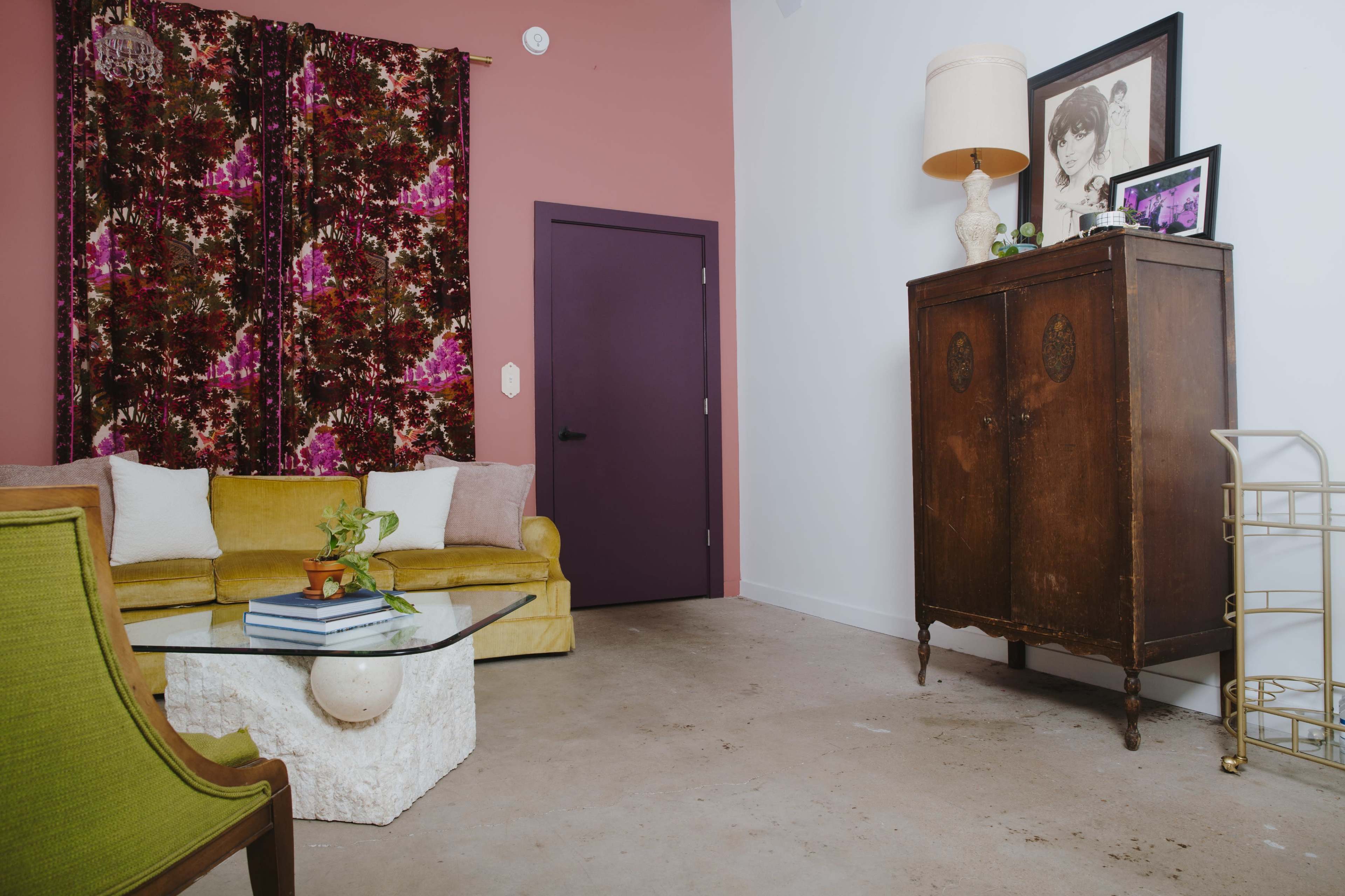 A living room features a yellow couch, a glass coffee table, an antique cabinet, and a wall adorned with floral curtains.
