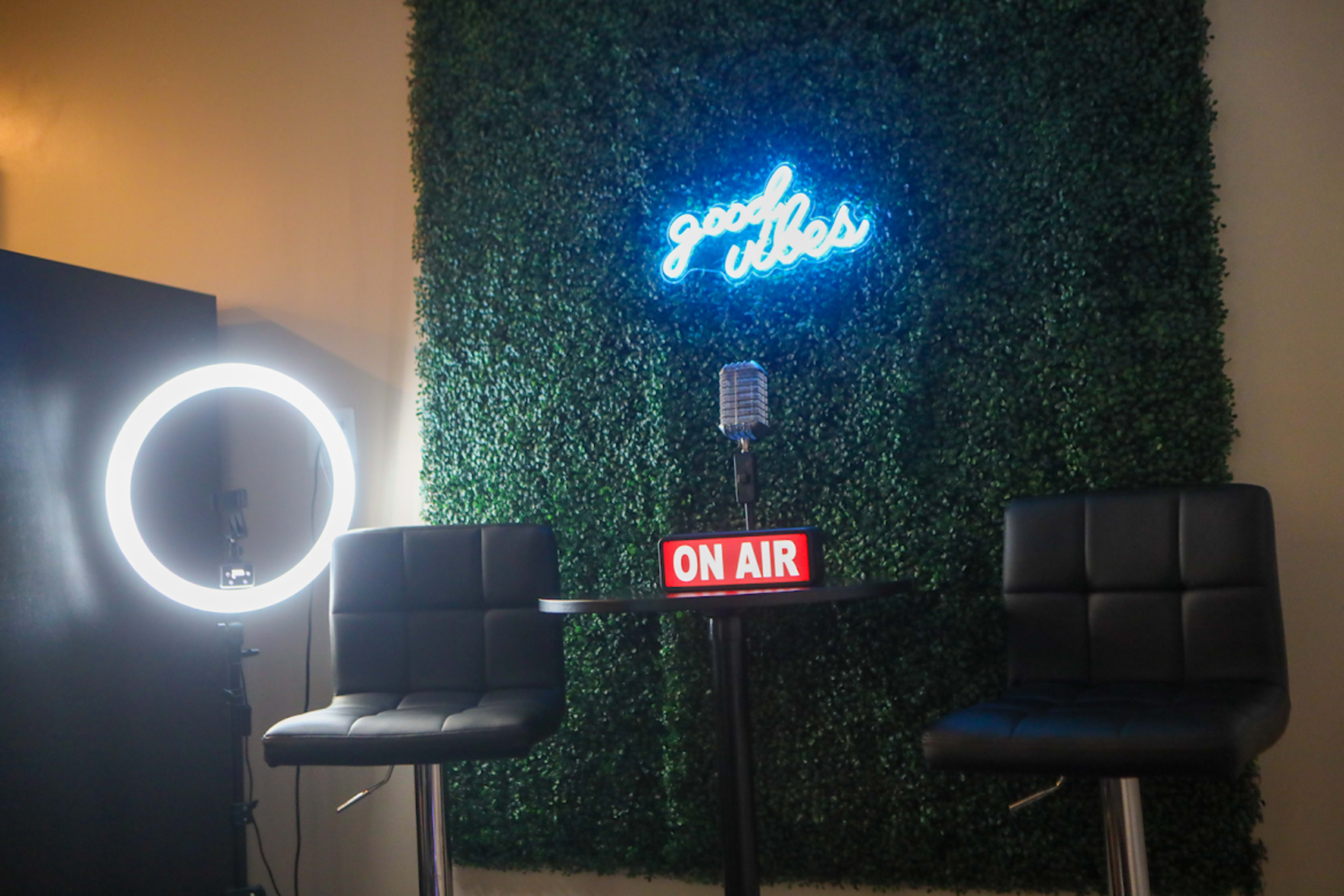 A small studio setup featuring two black chairs, a round table with an "ON AIR" sign, a microphone, and a neon sign that reads "good vibes" against a green foliage wall with a ring light.