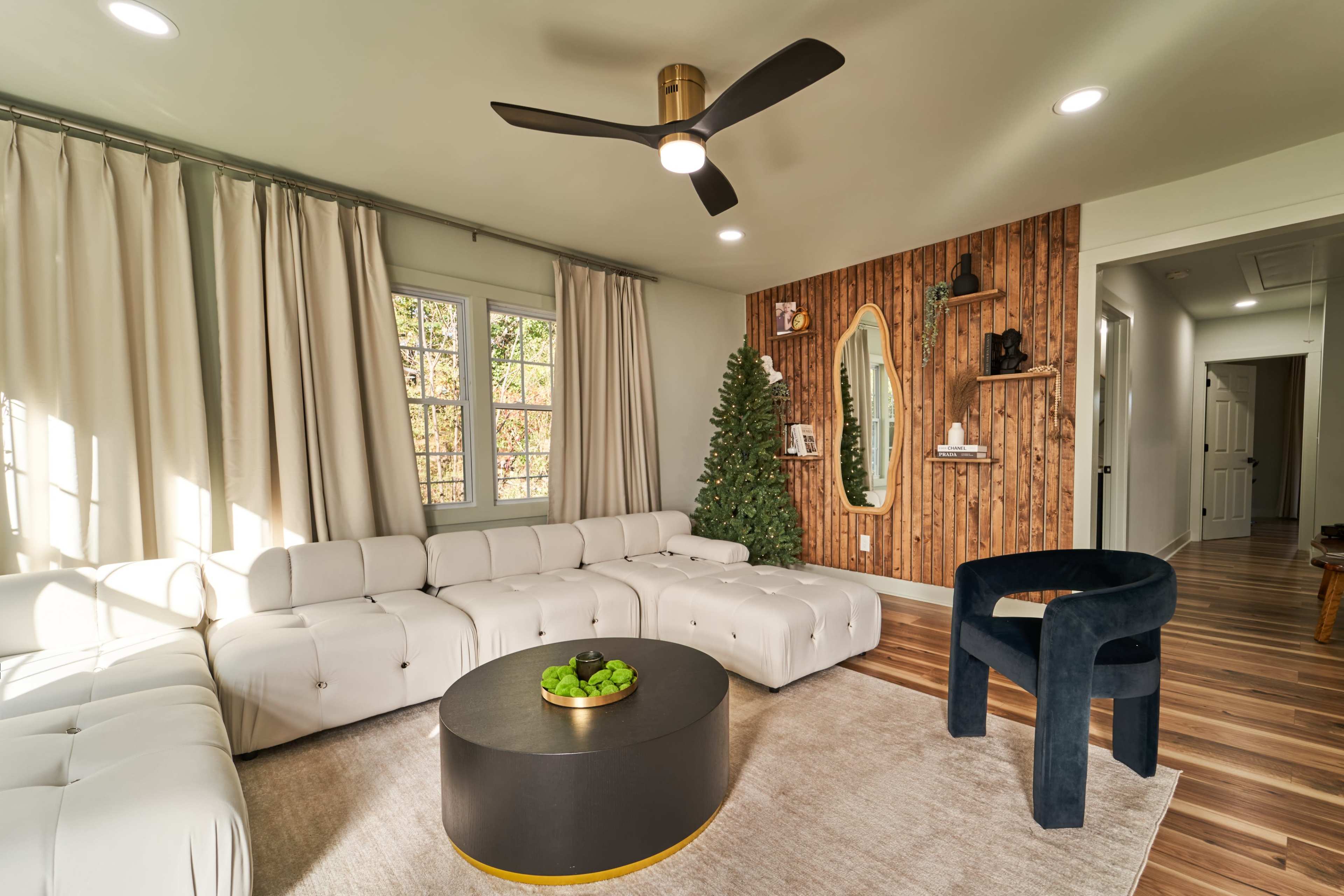 A modern living room featuring a white sectional sofa, a round black coffee table with green decor, a dark blue accent chair, and large windows with light-colored curtains.