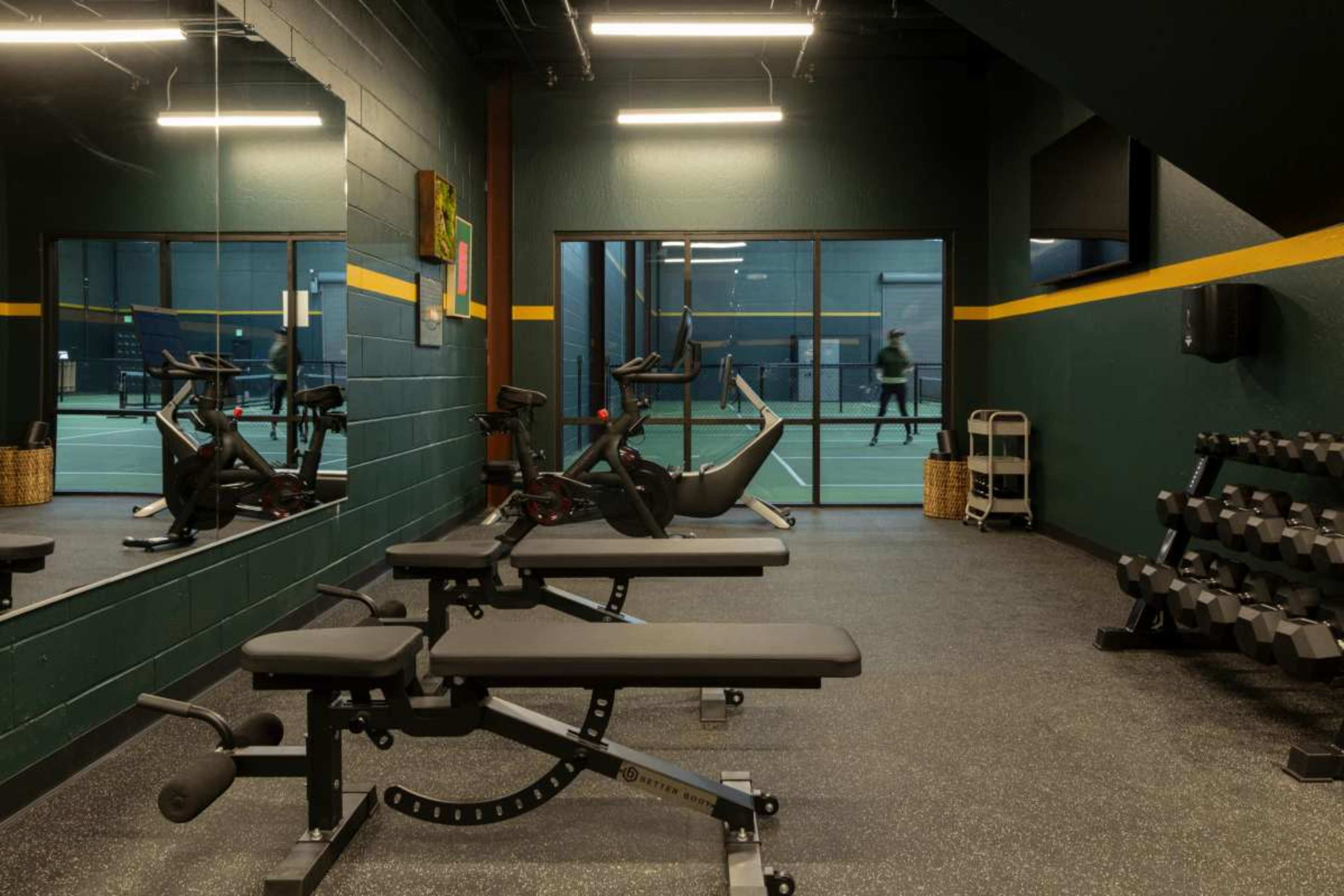 The image depicts a fitness room featuring exercise benches and dumbbells, with a view of a tennis court through large windows.