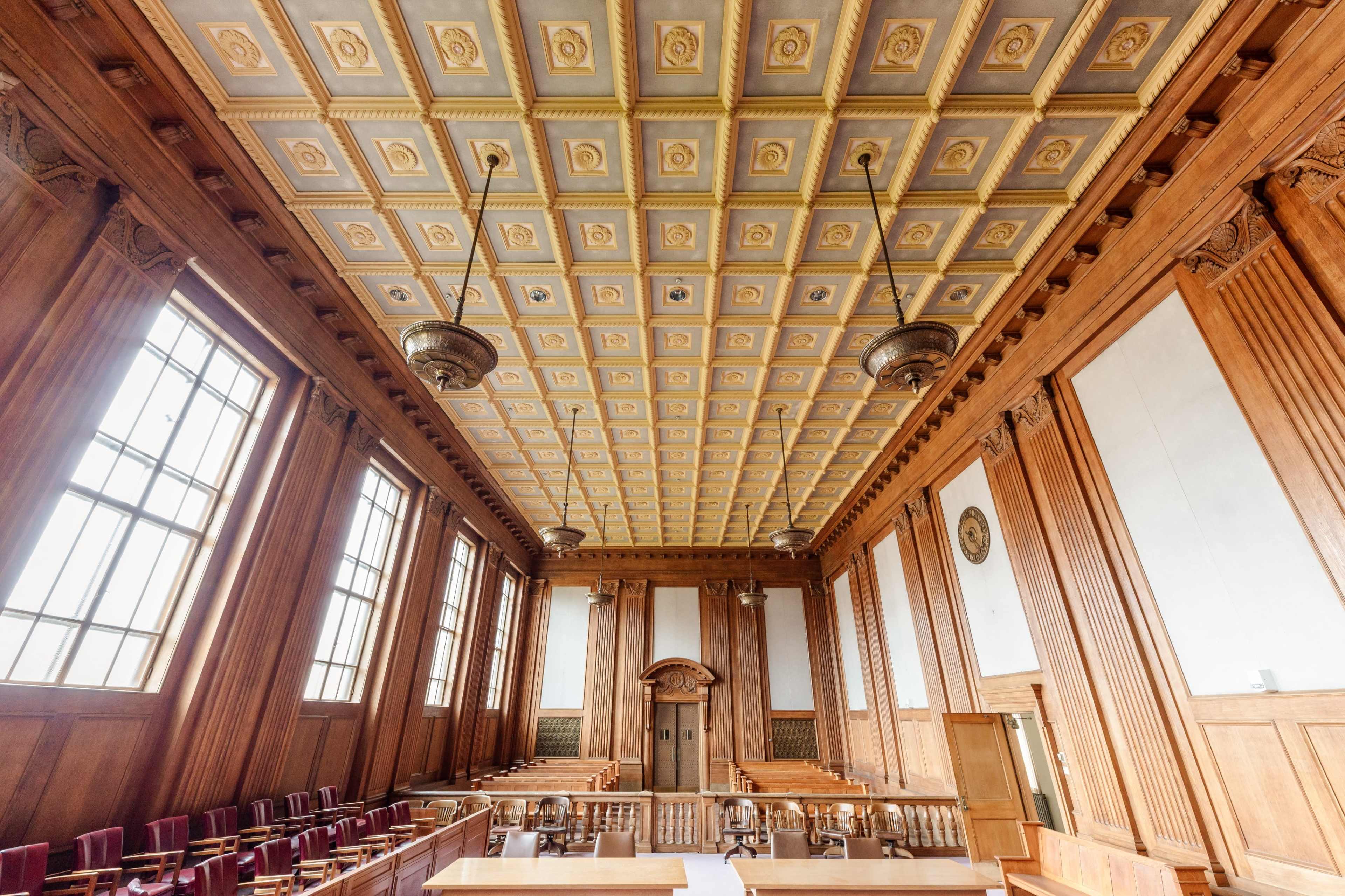 The image shows an ornate courtroom with a high, coffered ceiling, wooden paneling, and rows of wooden seating.