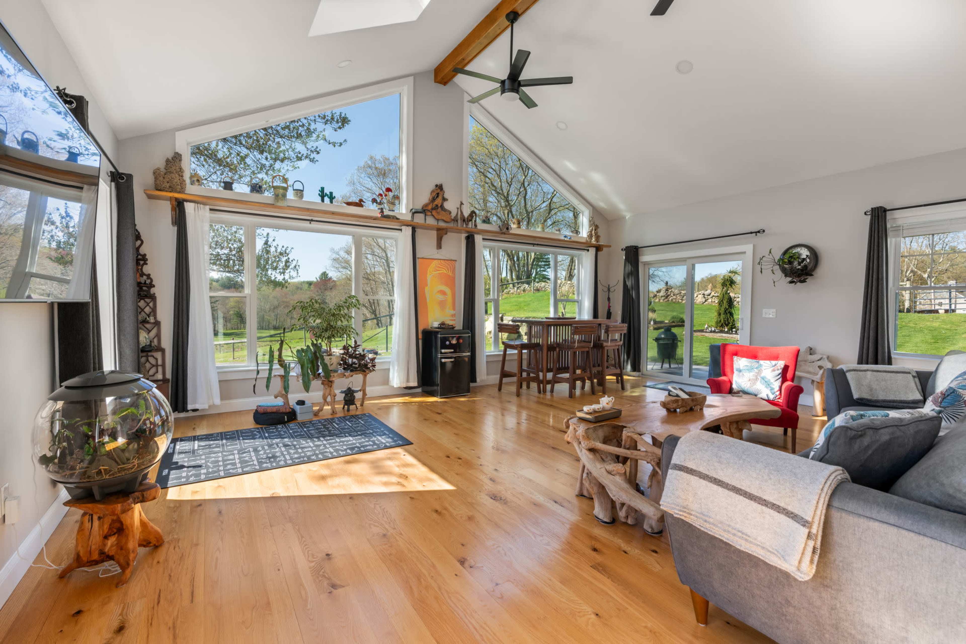 A spacious living room with large windows, wooden flooring, and modern furnishings, including a television and a fish tank.