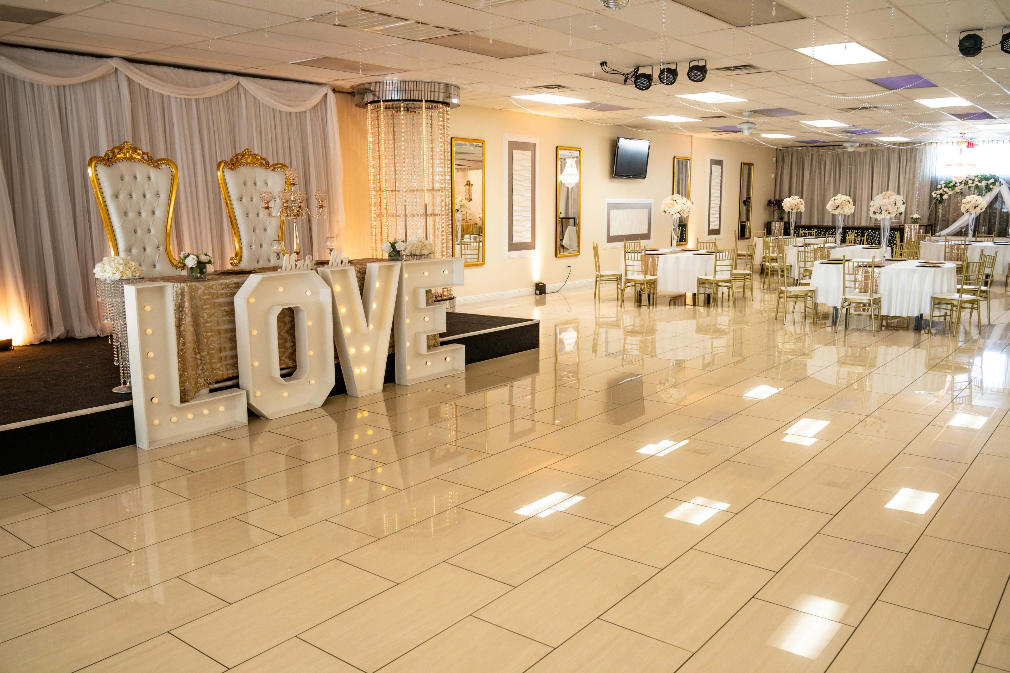 The image shows a decorated event space featuring large illuminated letters spelling "LOVE," elegant chairs on a stage, and round dining tables set up for guests.