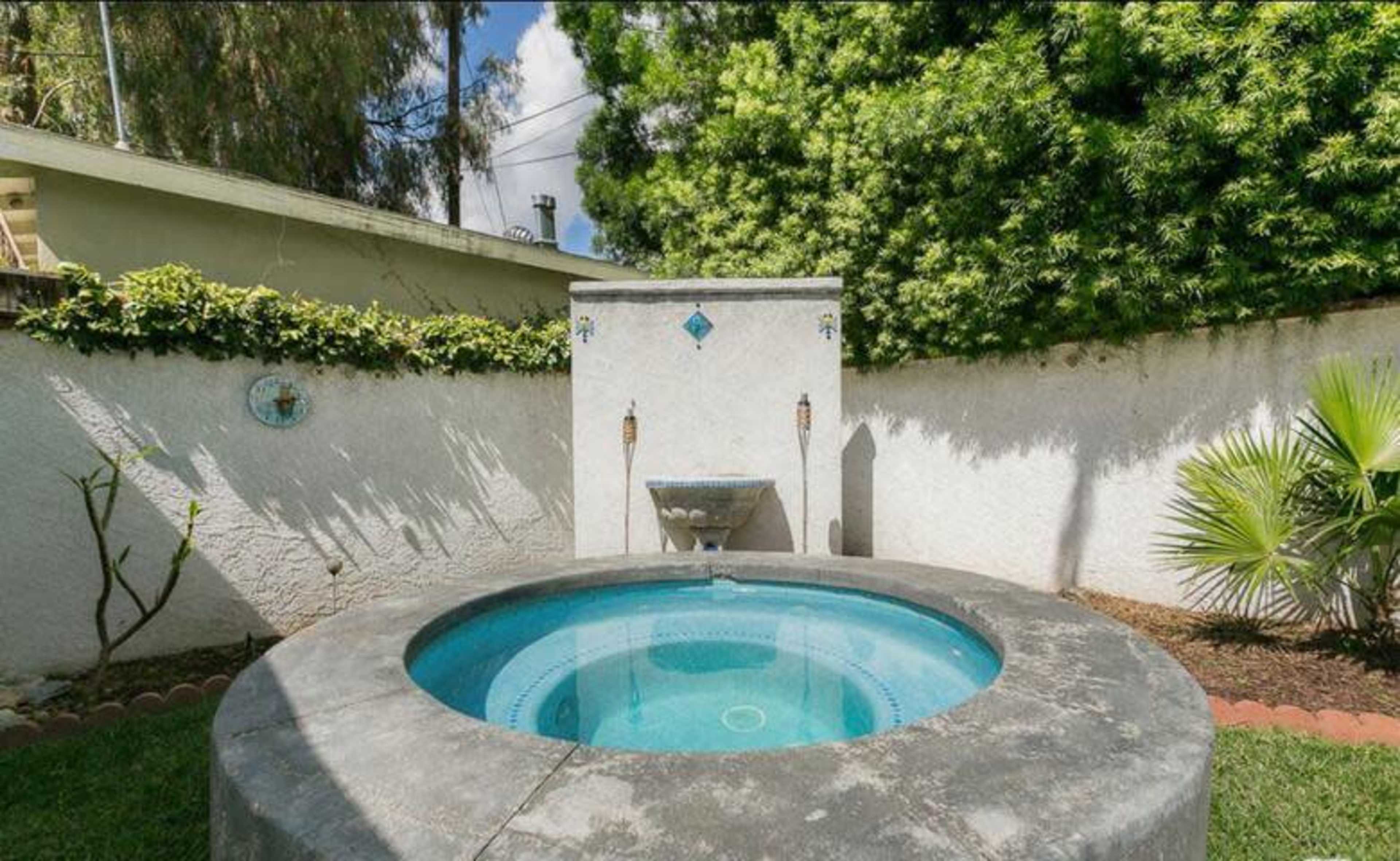 The image shows a circular hot tub surrounded by a landscaped area, with a stone wall and a water fountain in the background.