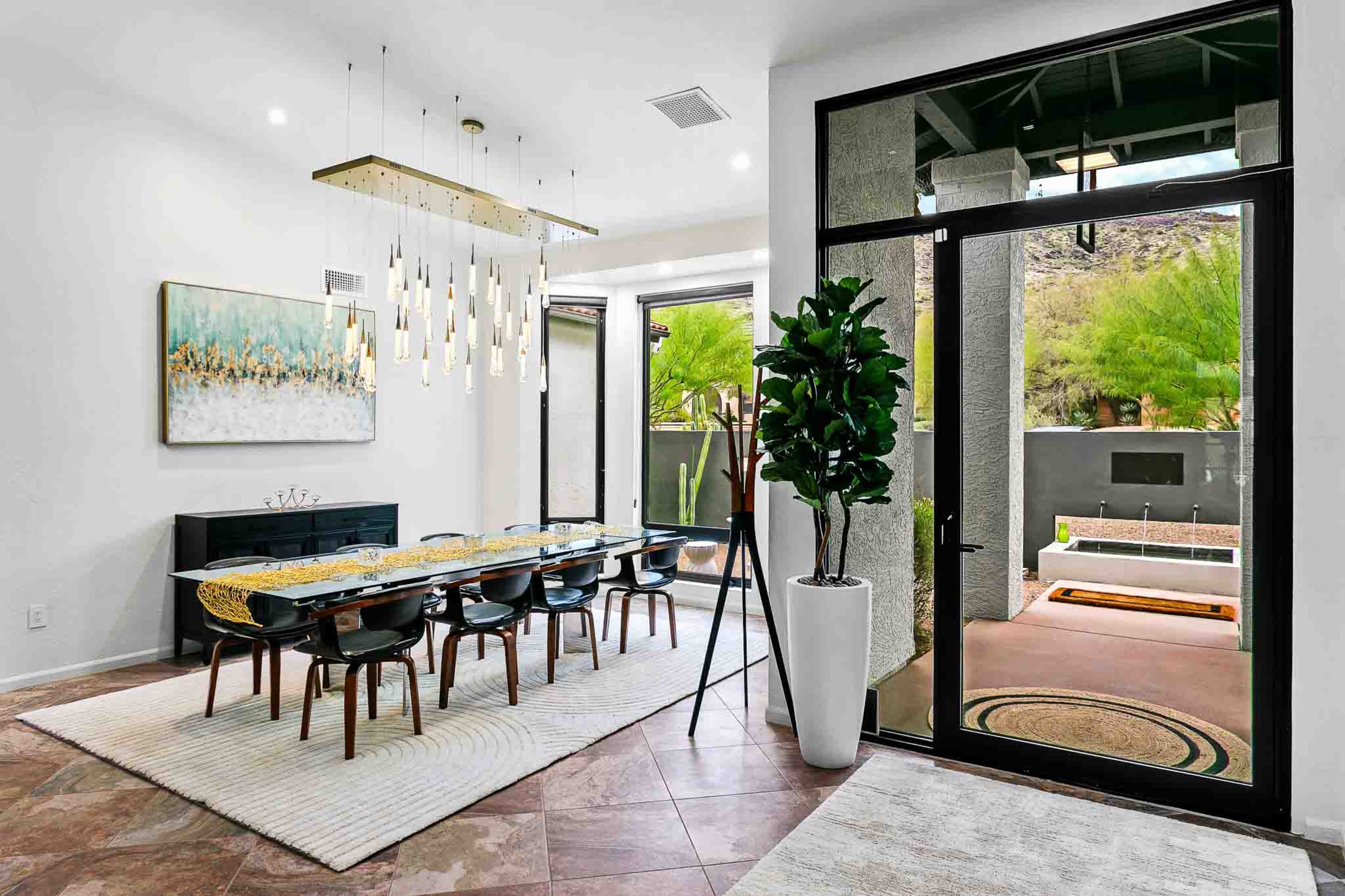 A modern dining area features a long table surrounded by chairs, a decorative light fixture above, and large windows that open to an outdoor space.