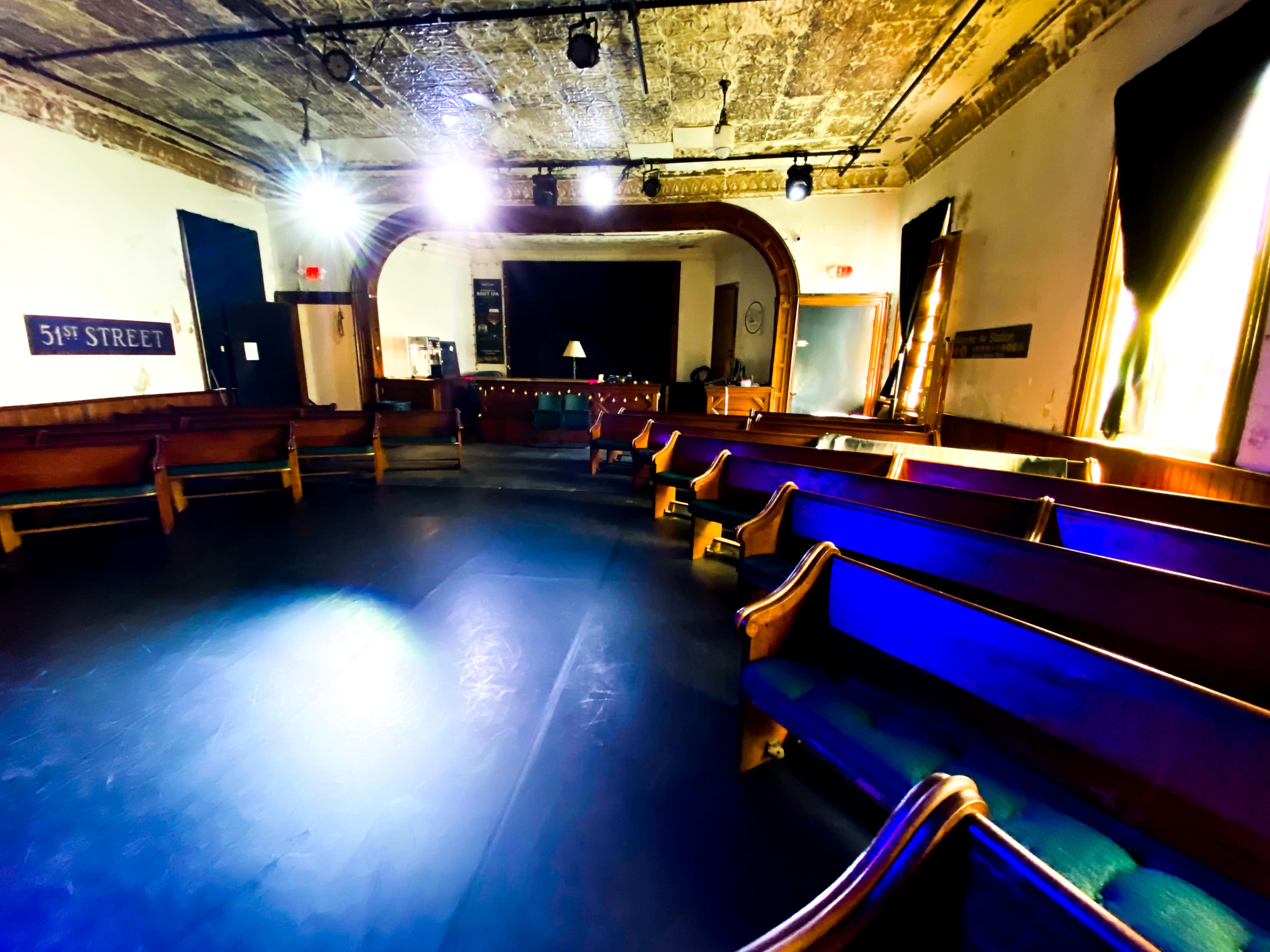 The image shows an empty, circular space with wooden benches arranged around a central area, illuminated by stage lights and featuring a backdrop with a sign reading "5TH STREET."