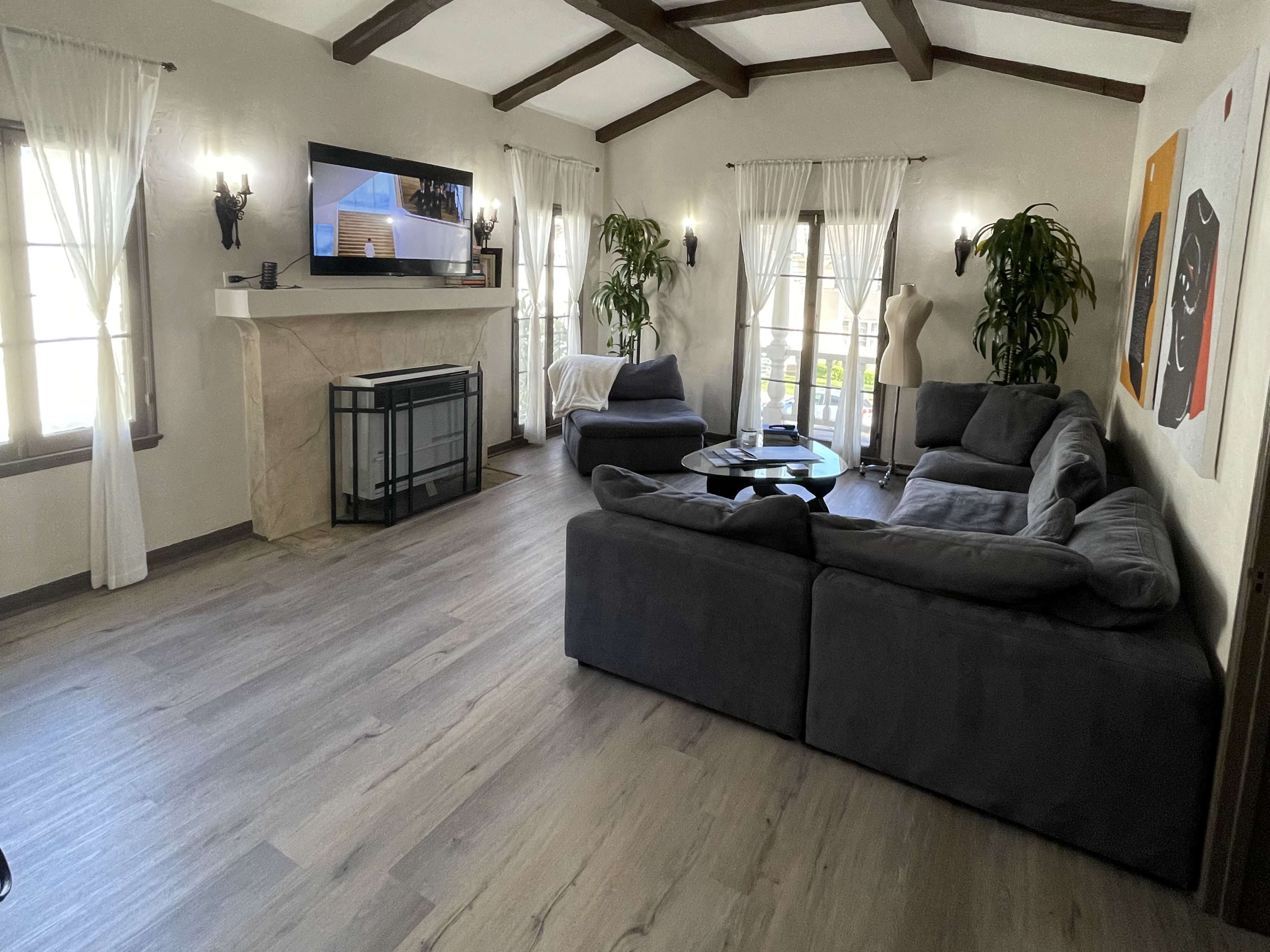 The living room features a gray sectional sofa, a coffee table, and a television mounted on a light-colored stone mantel, with large windows allowing natural light to fill the space.