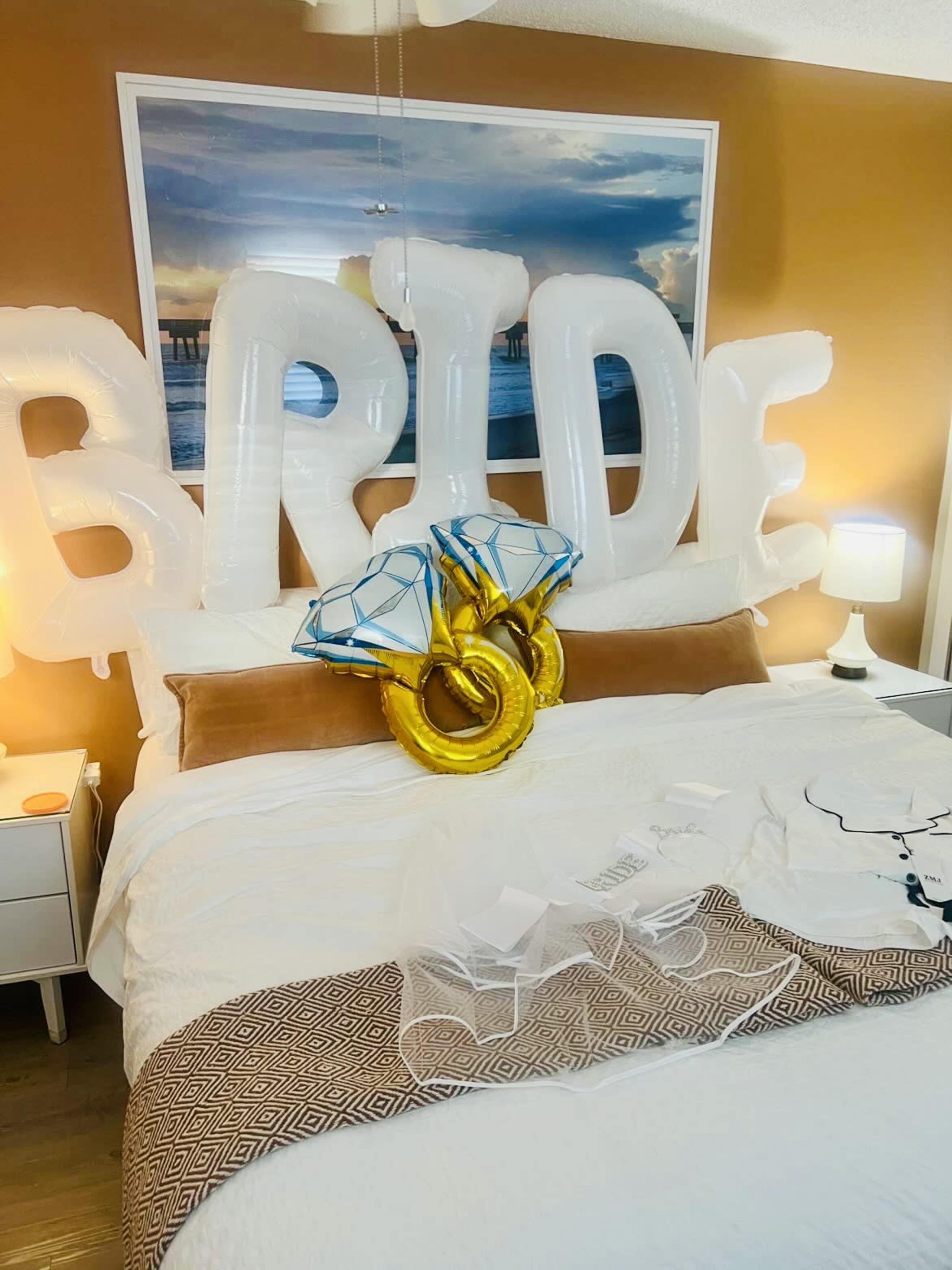 The image shows a bedroom with large inflatable letters spelling "BRIDE," two diamond-shaped balloons, and a white dress laid out on the bed.