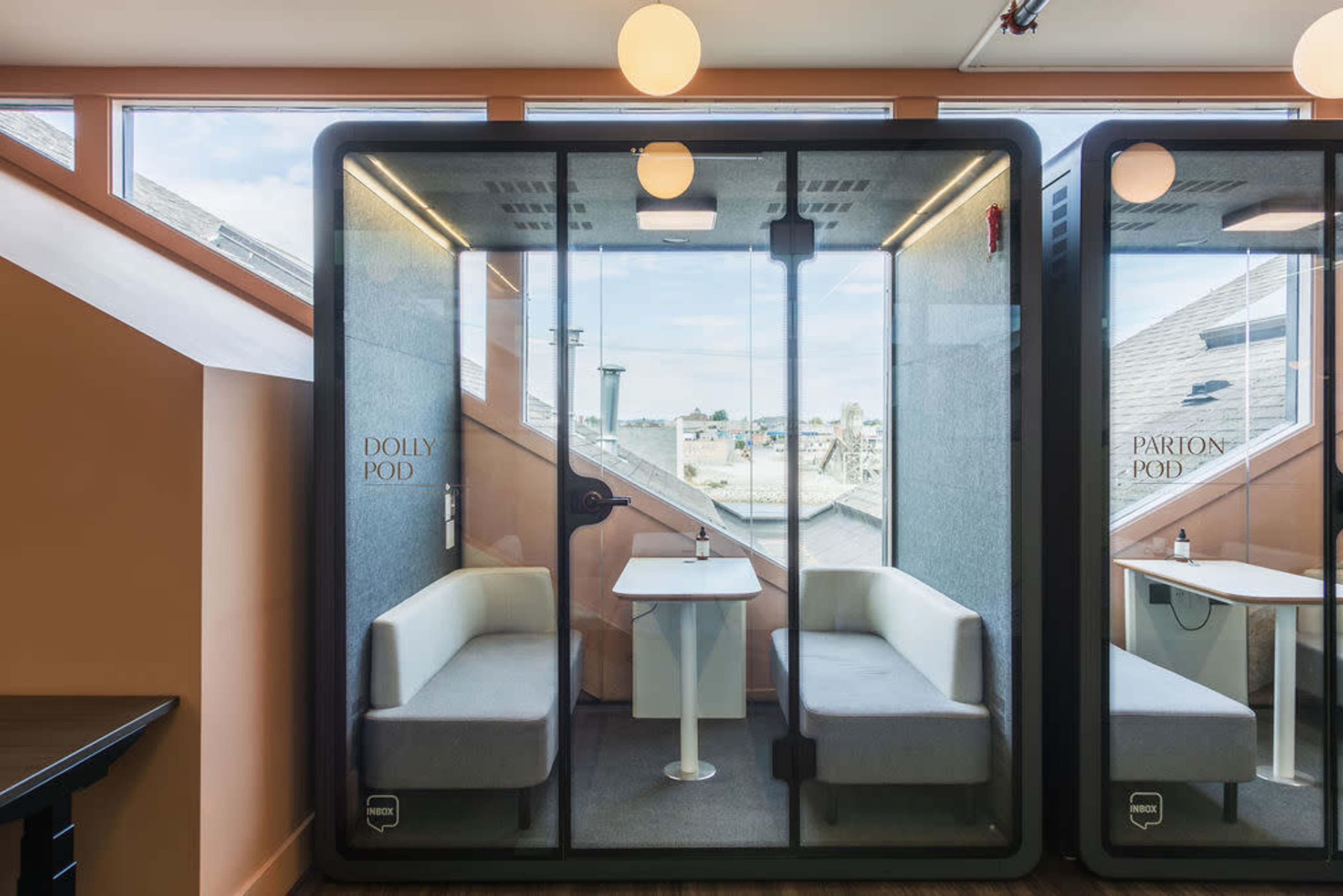 The image shows two soundproof meeting pods with glass doors, furnished with a table and seating, located in a modern office environment.