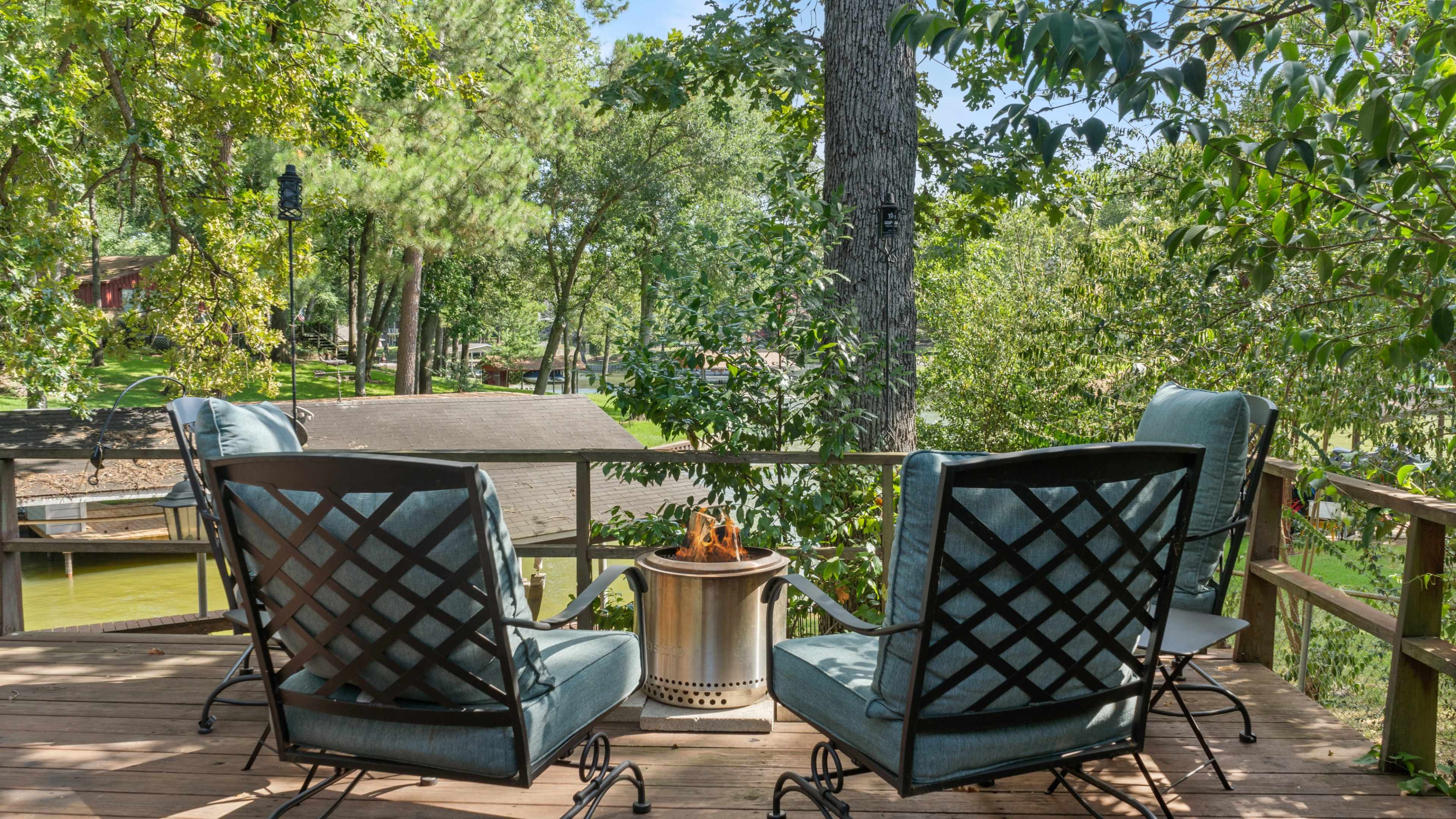 A wooden deck features two metal chairs facing a small fire pit, surrounded by trees and a view of a lake.