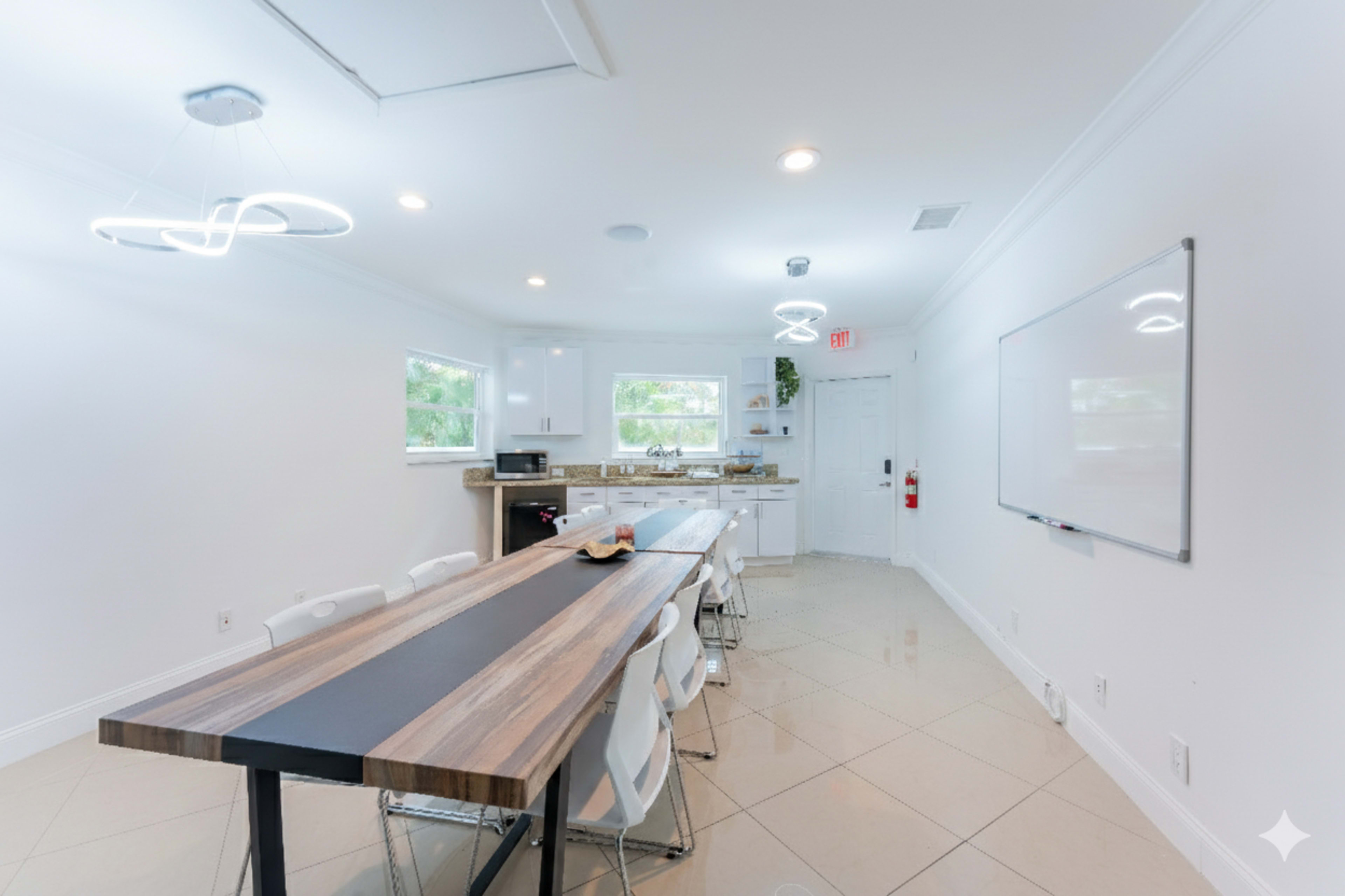 A bright, modern conference room features a long table surrounded by white chairs, with a kitchenette and whiteboard visible in the background.