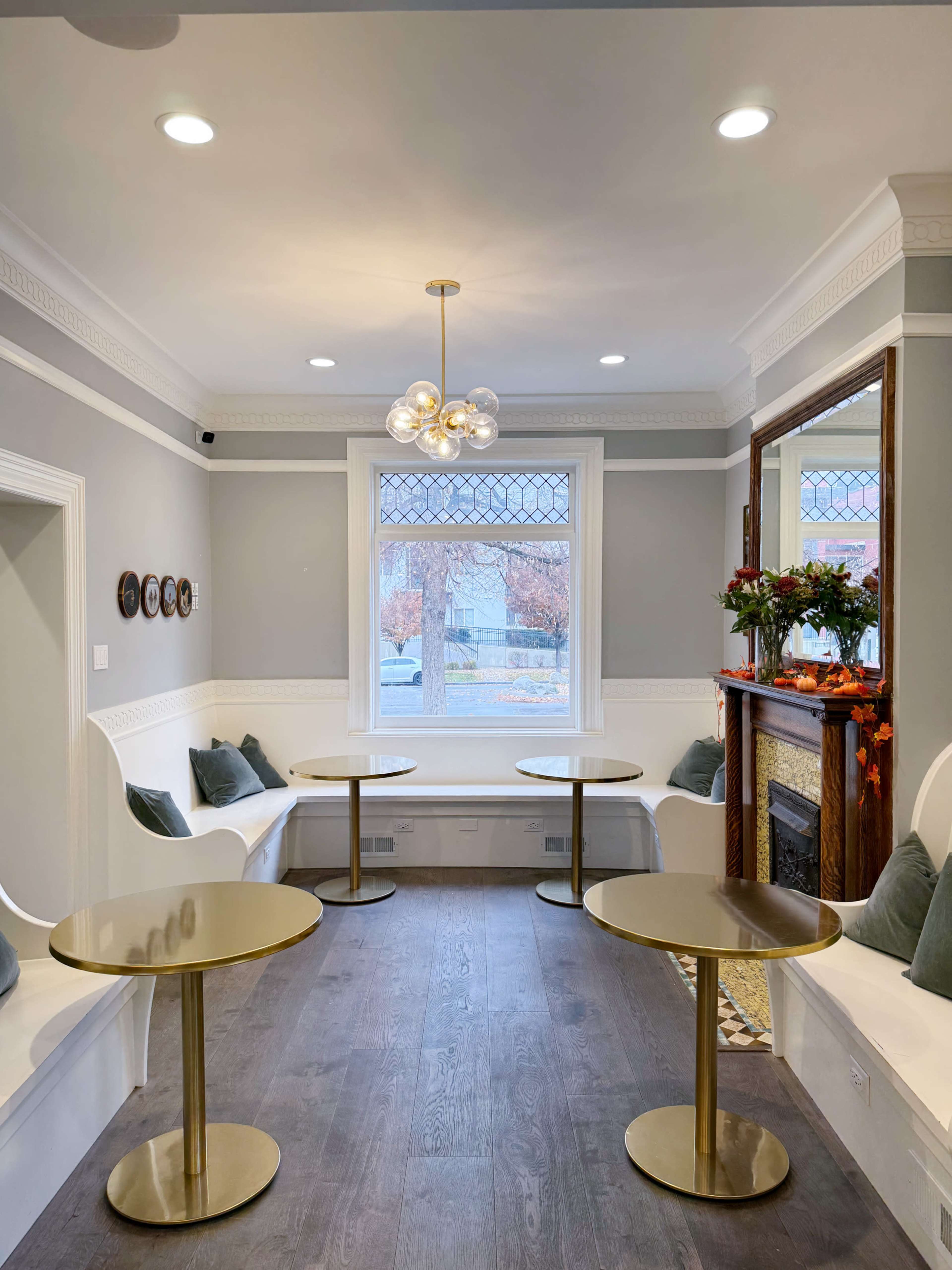A cozy corner seating area with two round gold tables, a decorative fireplace, and a large window framed by light-colored walls.