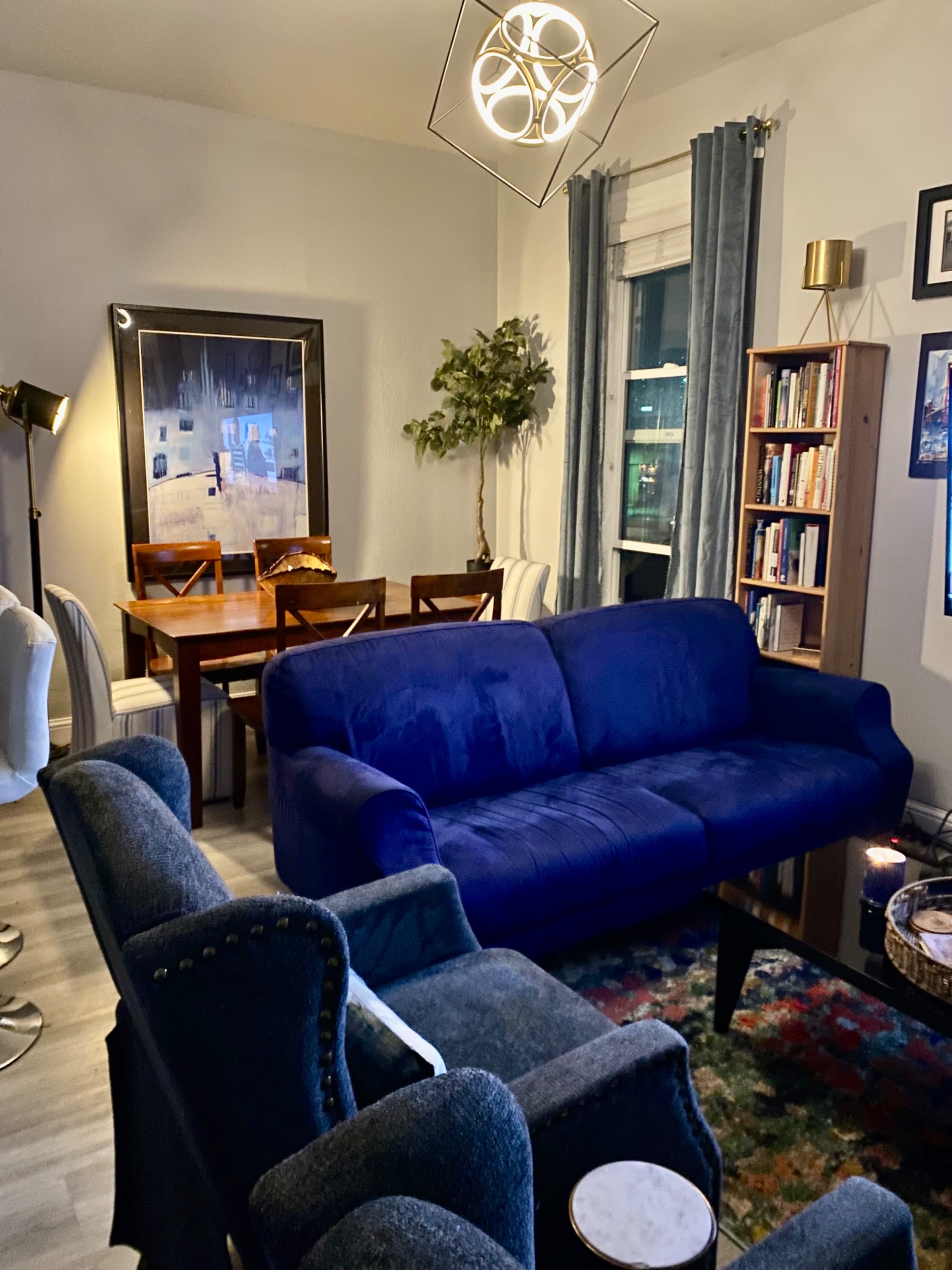 The image shows a living room featuring a blue couch, two armchairs, a dining table with chairs, a bookshelf, and a decorative pendant light.