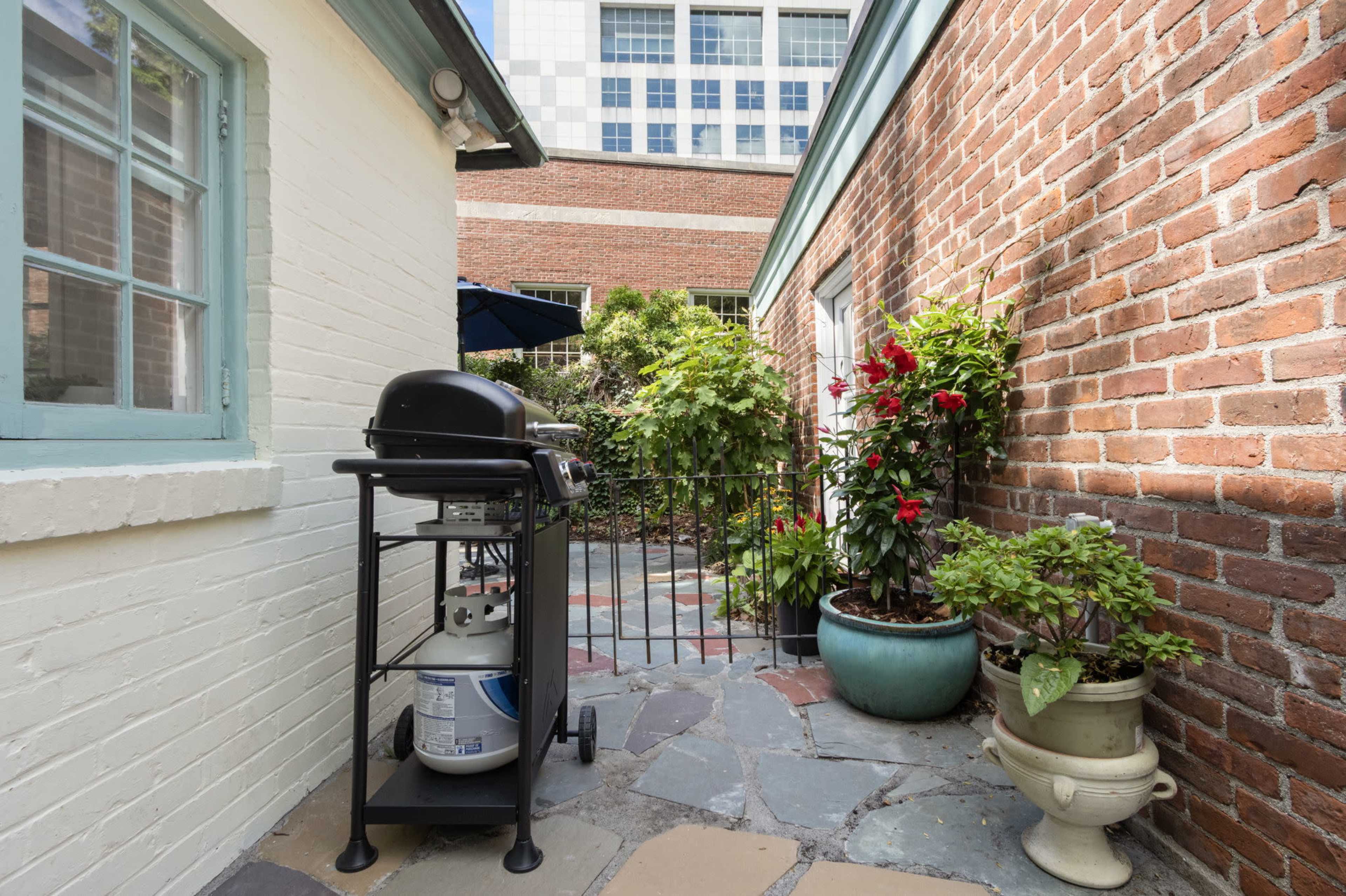 A black gas grill is positioned on a stone patio surrounded by potted plants and red flowers, with a brick wall and a building visible in the background.