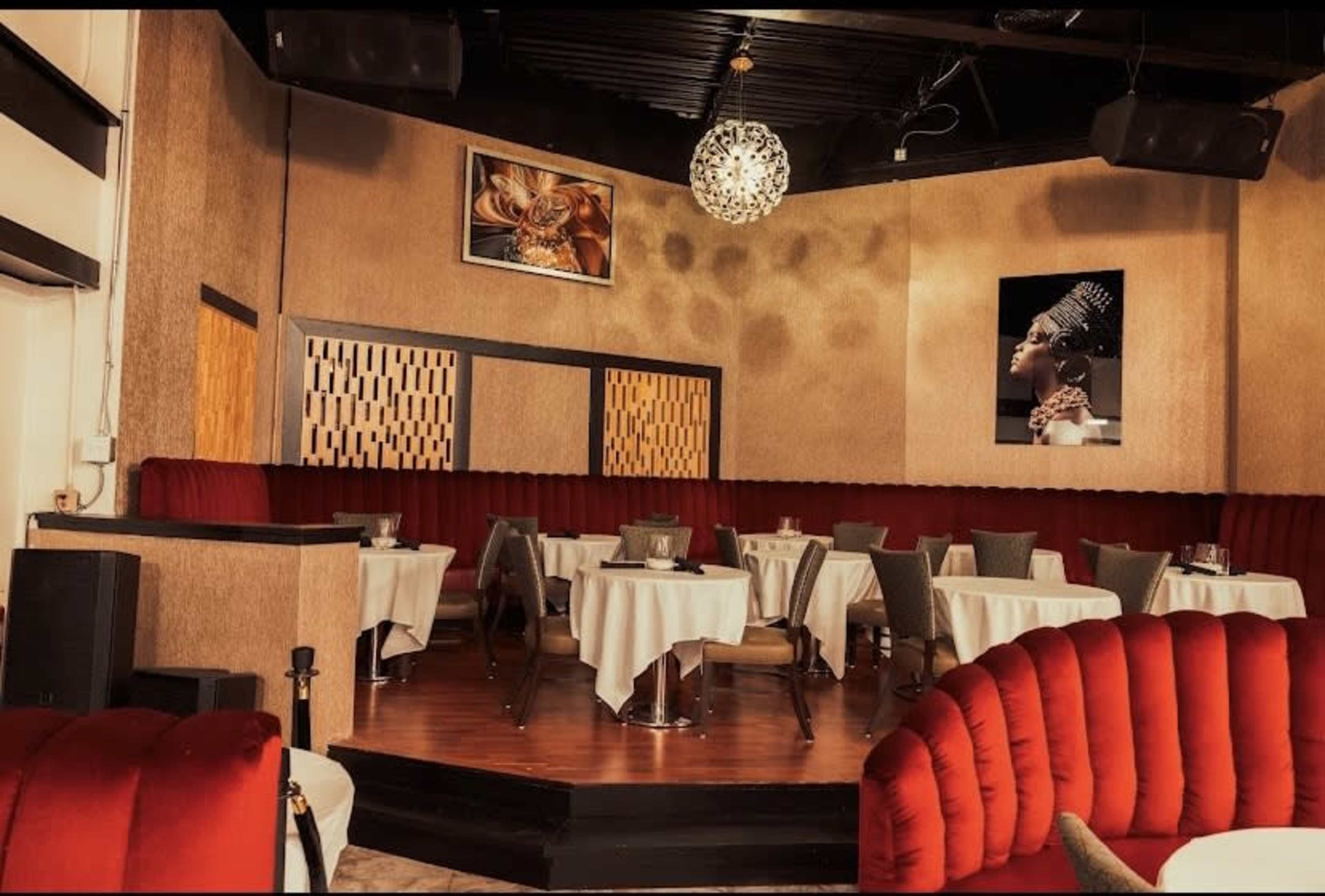 The image shows a stylish restaurant interior featuring round tables covered with white tablecloths, red upholstered seating, and decorative artwork on the walls.