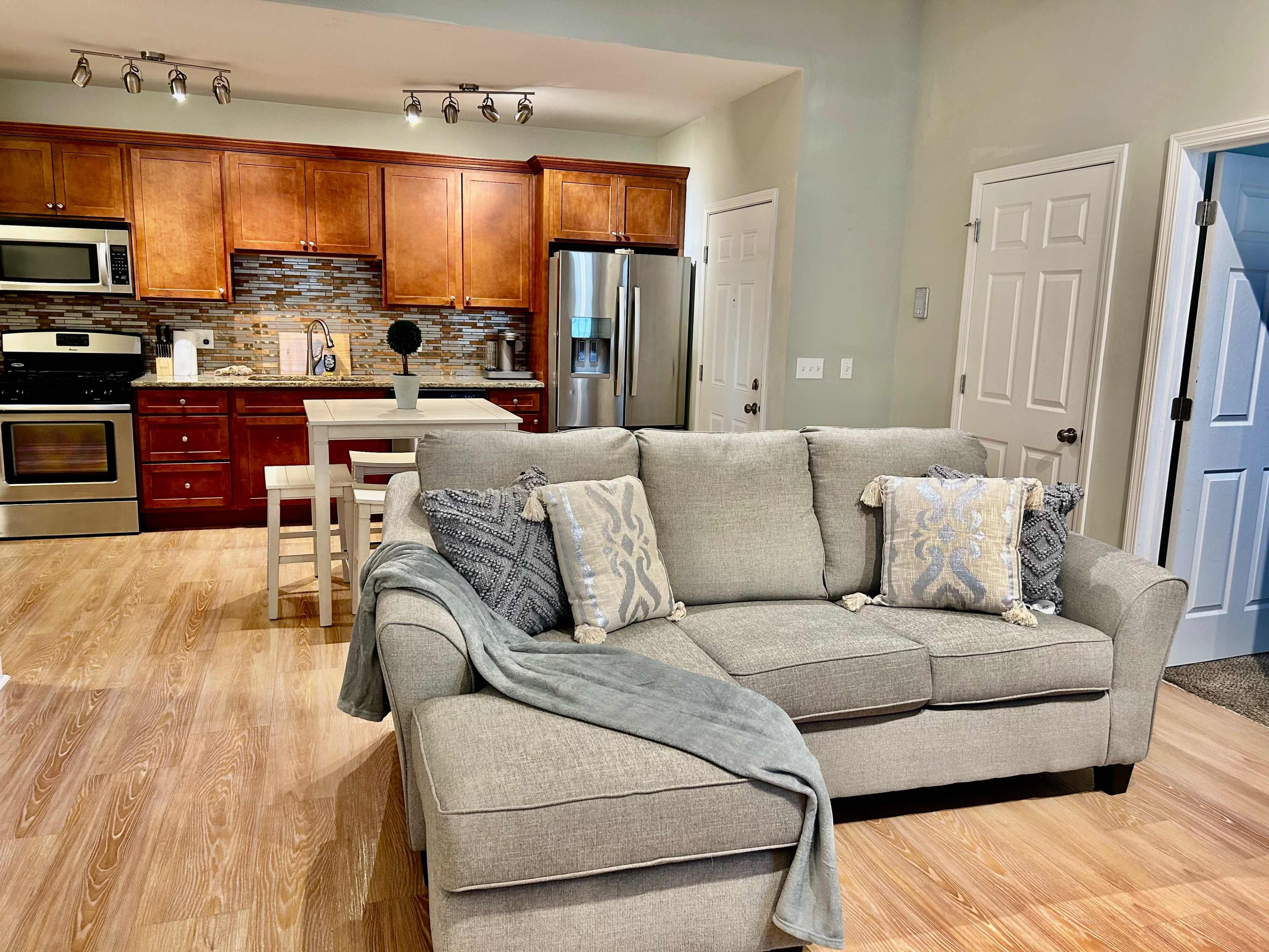 A living area with a gray couch adorned with decorative pillows, a wooden kitchen in the background featuring stainless steel appliances, and a dining table with chairs.