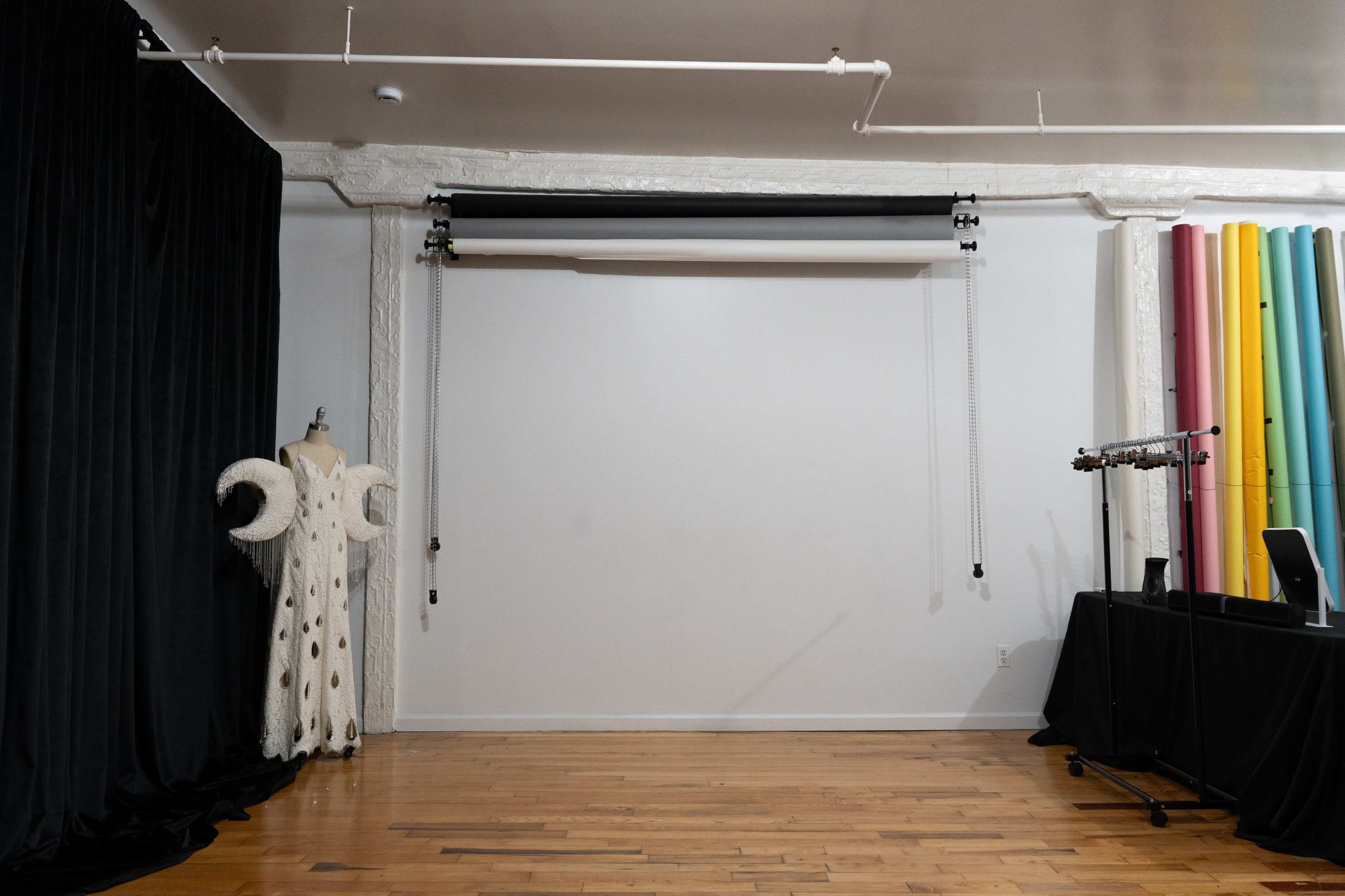 The image shows a studio space with a blank wall, a garment on a mannequin, and a backdrop support system on the ceiling, alongside a table with color rolls in the background.