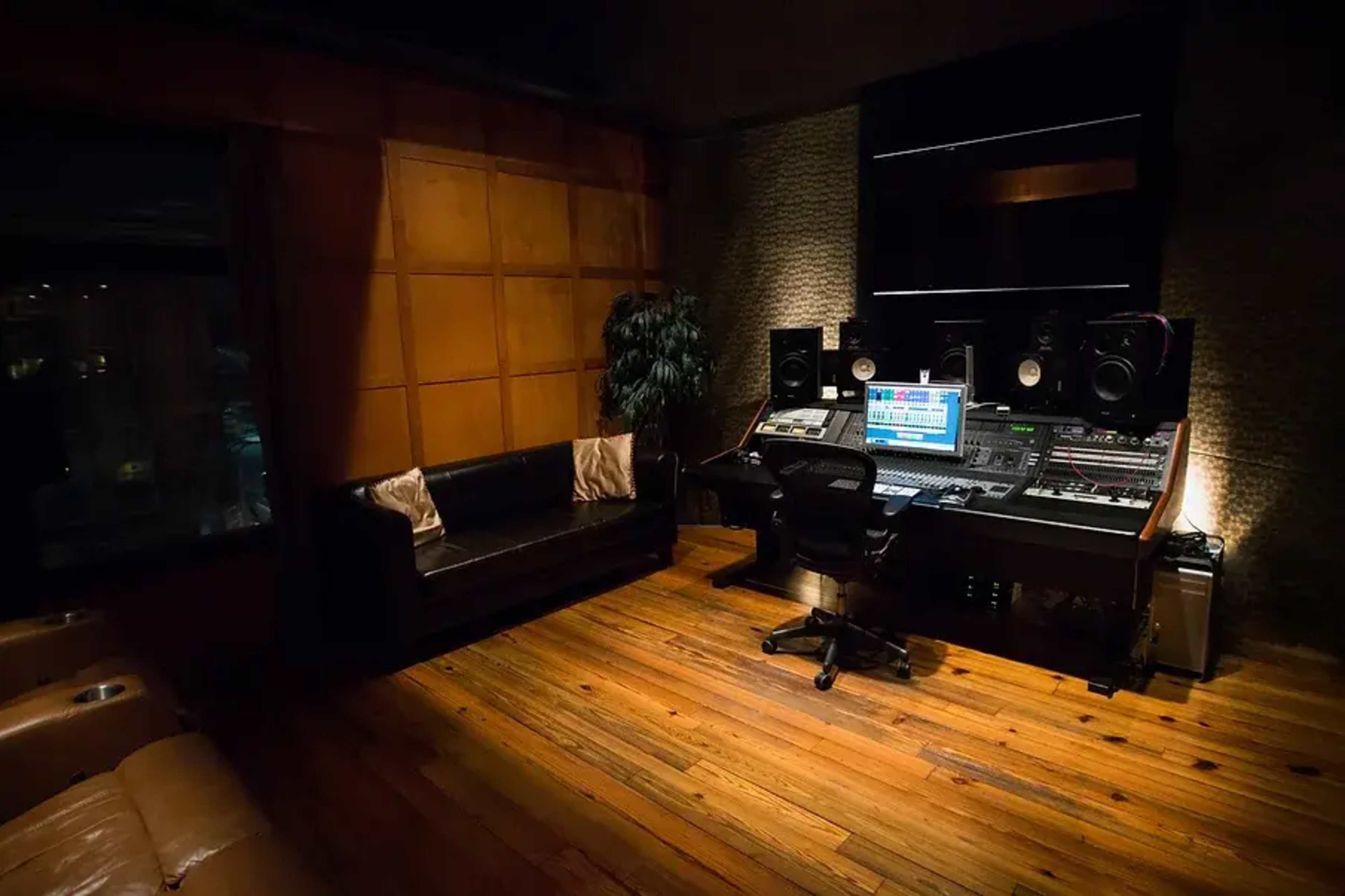 A music recording studio with a central mixing console, a black ergonomic chair, and a leather couch against a wooden wall.
