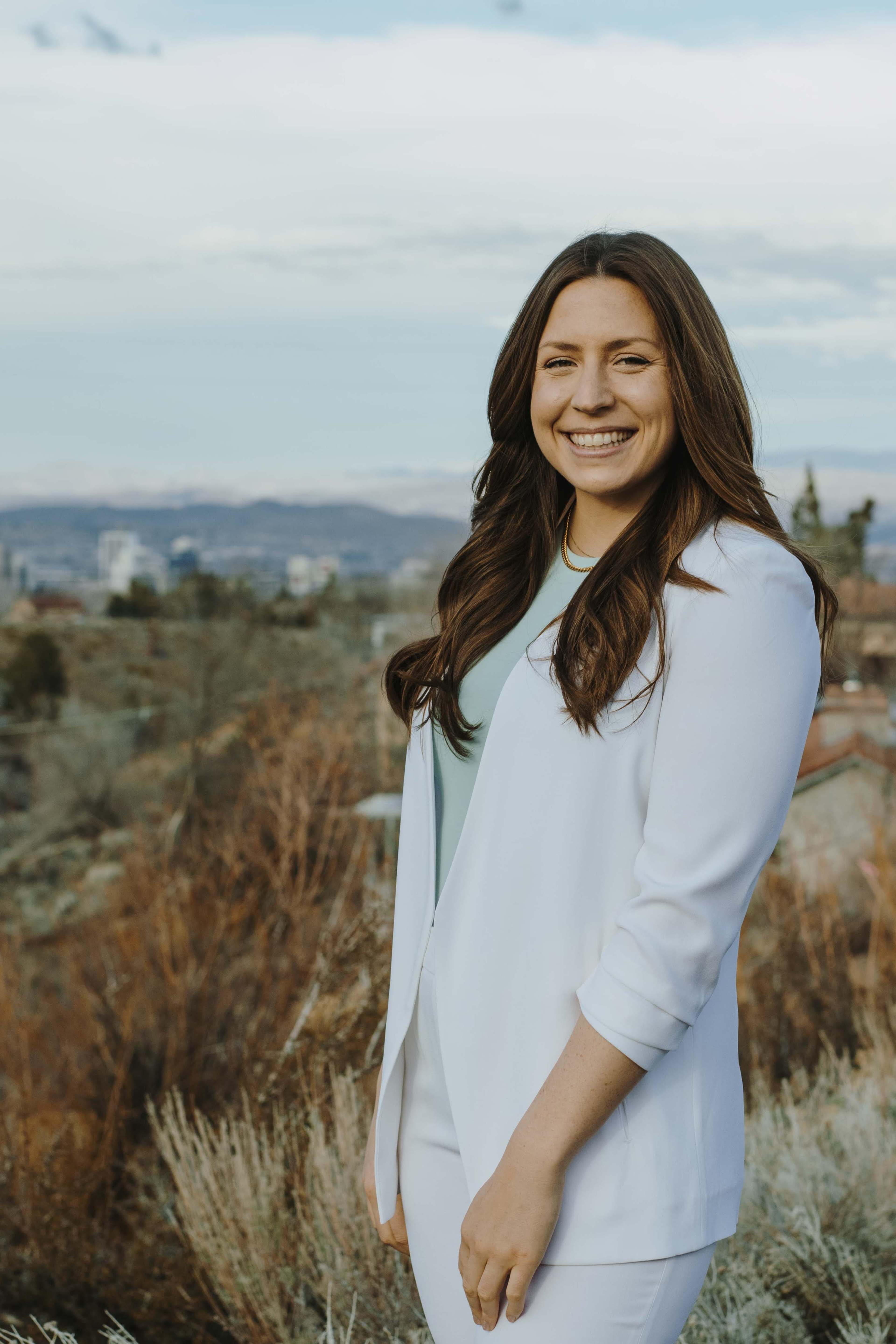 A woman in a white blazer stands outdoors with a landscape of hills and buildings in the background.