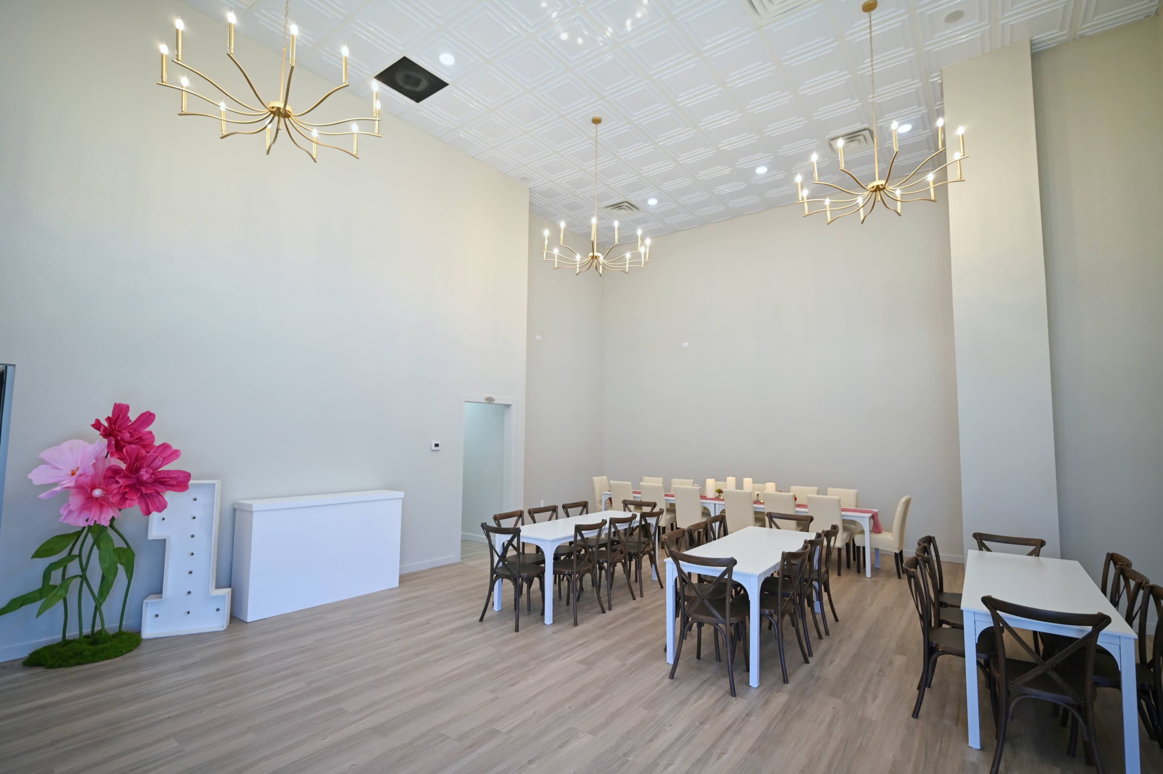 The image shows a spacious, well-lit room with several tables and chairs arranged around the center, featuring chandeliers on the ceiling and a large pink flower decoration at one side.
