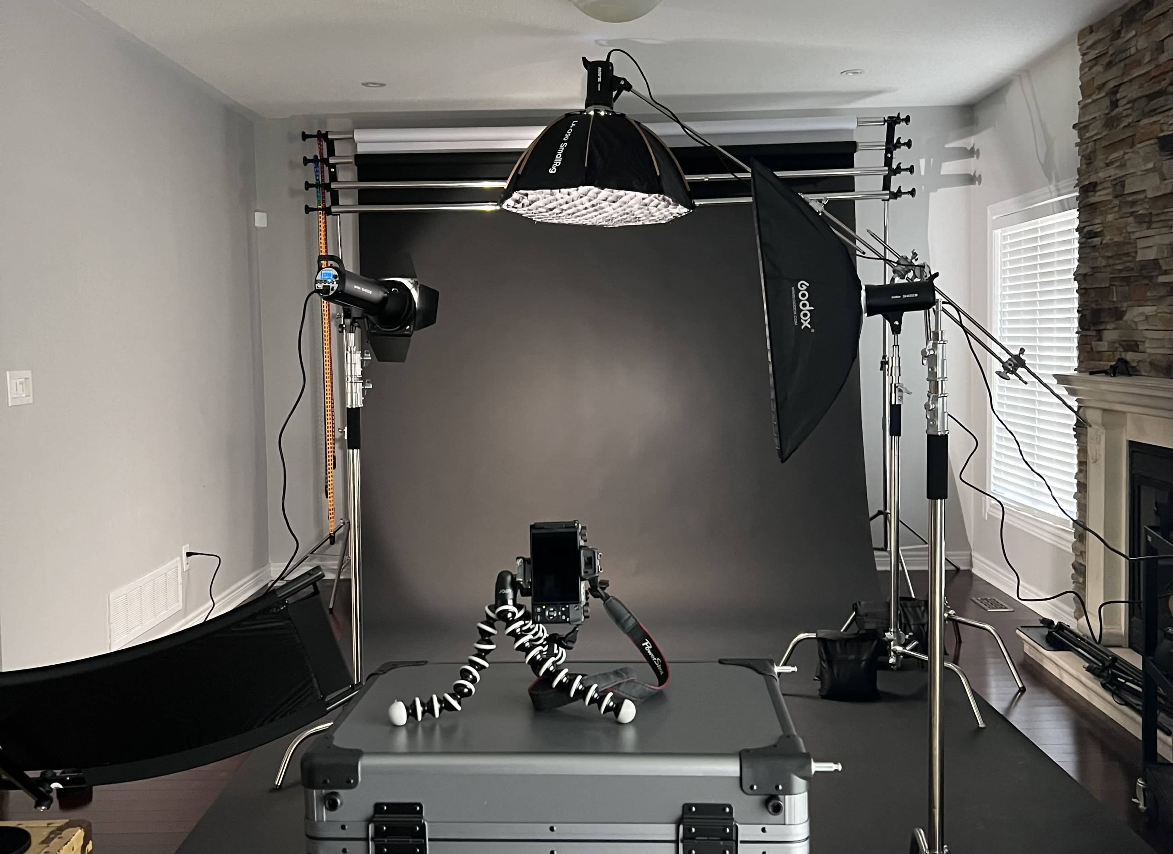 The image shows a photography studio setup with various lighting equipment, a camera on a tripod, and a dark backdrop.