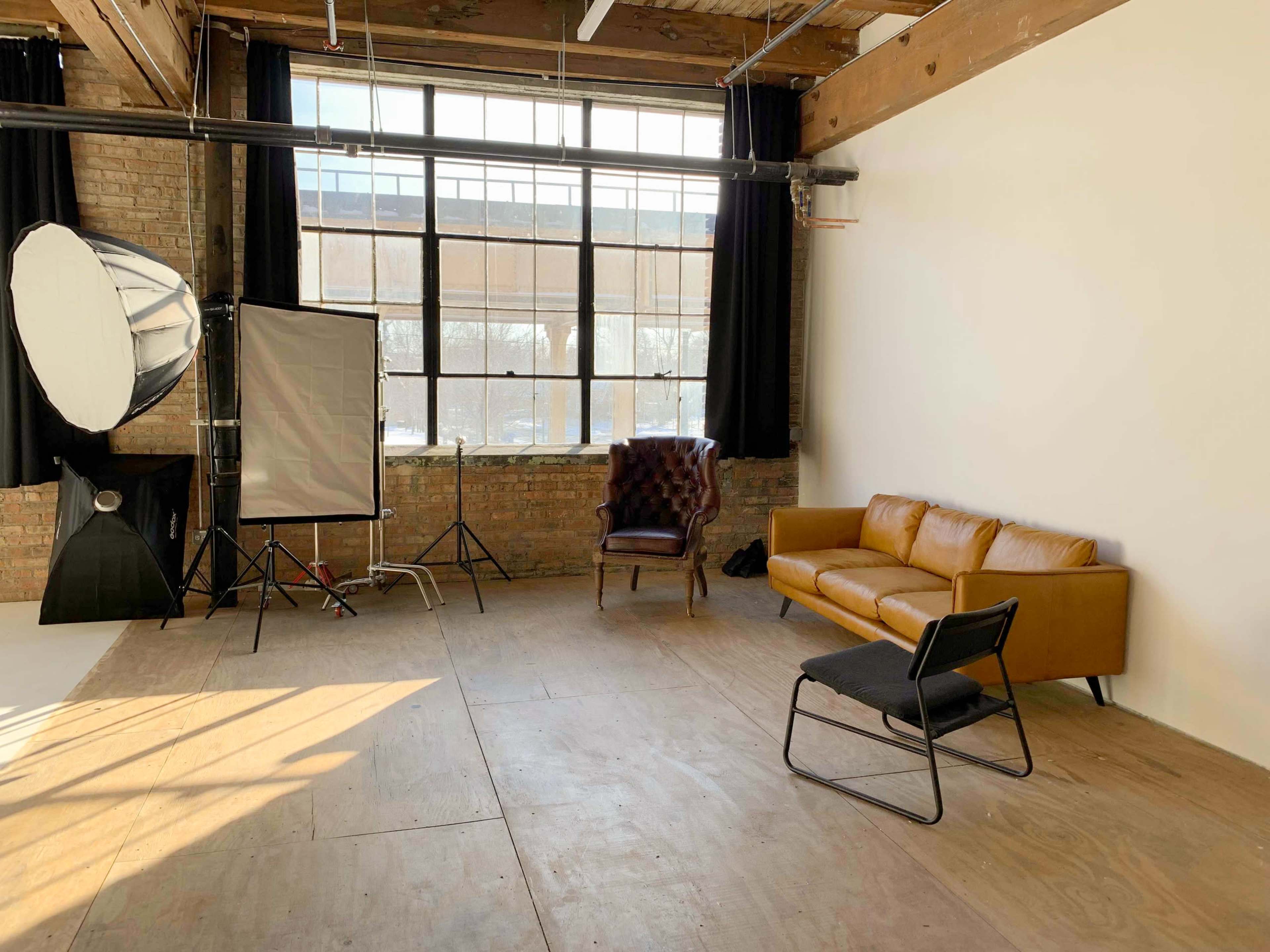 A spacious photography studio with large windows, a brown leather couch, a vintage armchair, and softbox lighting equipment.