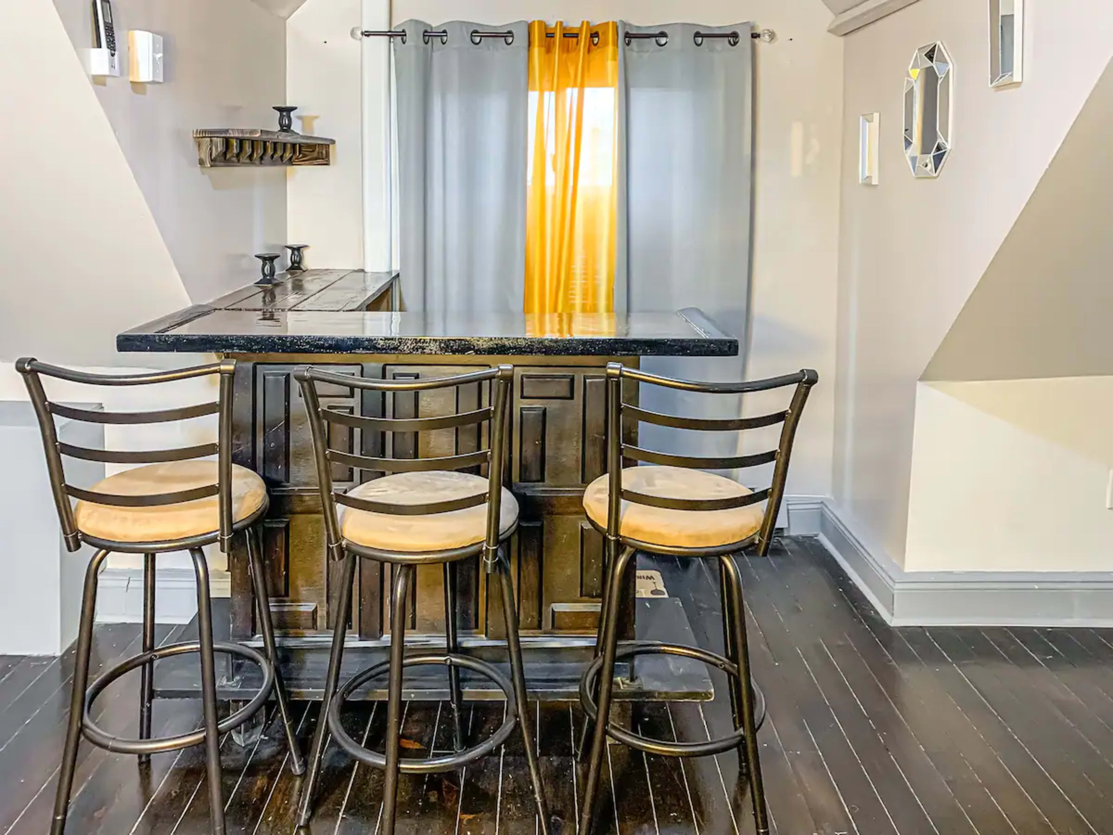 A small home bar with four black stools, a dark countertop, and a window dressed with yellow curtains.