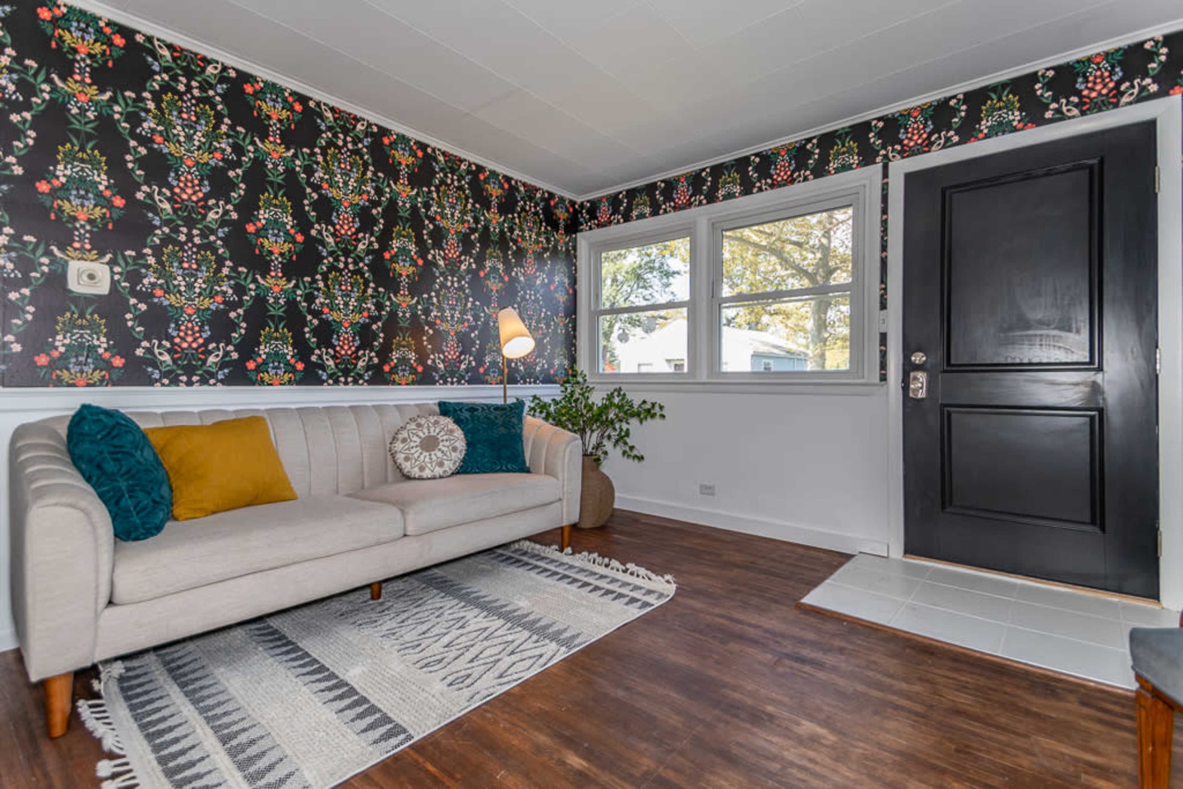 A cozy living room features a light-colored couch, a patterned area rug, and a door leading outside, surrounded by floral wallpaper.
