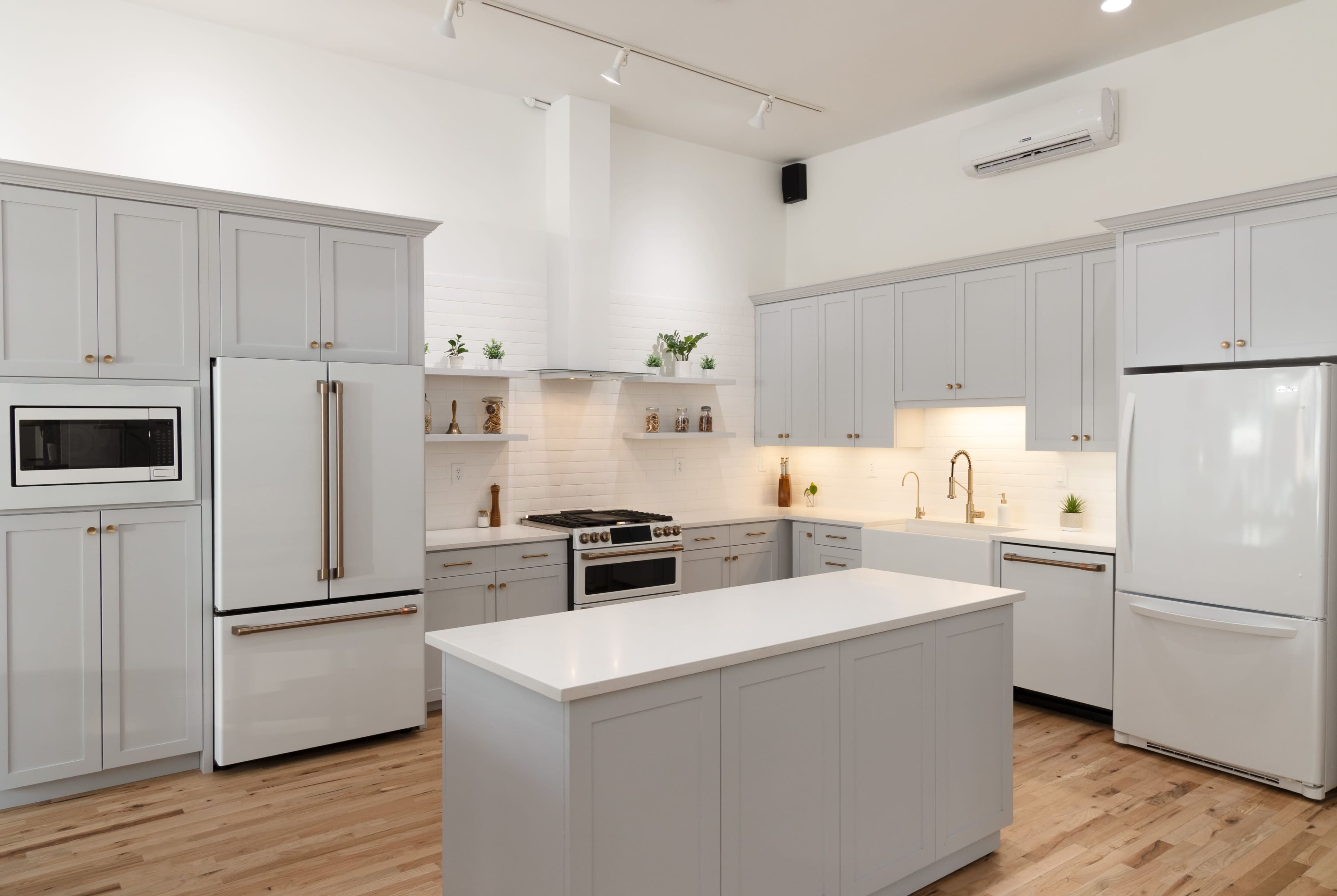 The kitchen features light gray cabinetry, a large central island, and stainless steel appliances against a bright, airy backdrop.