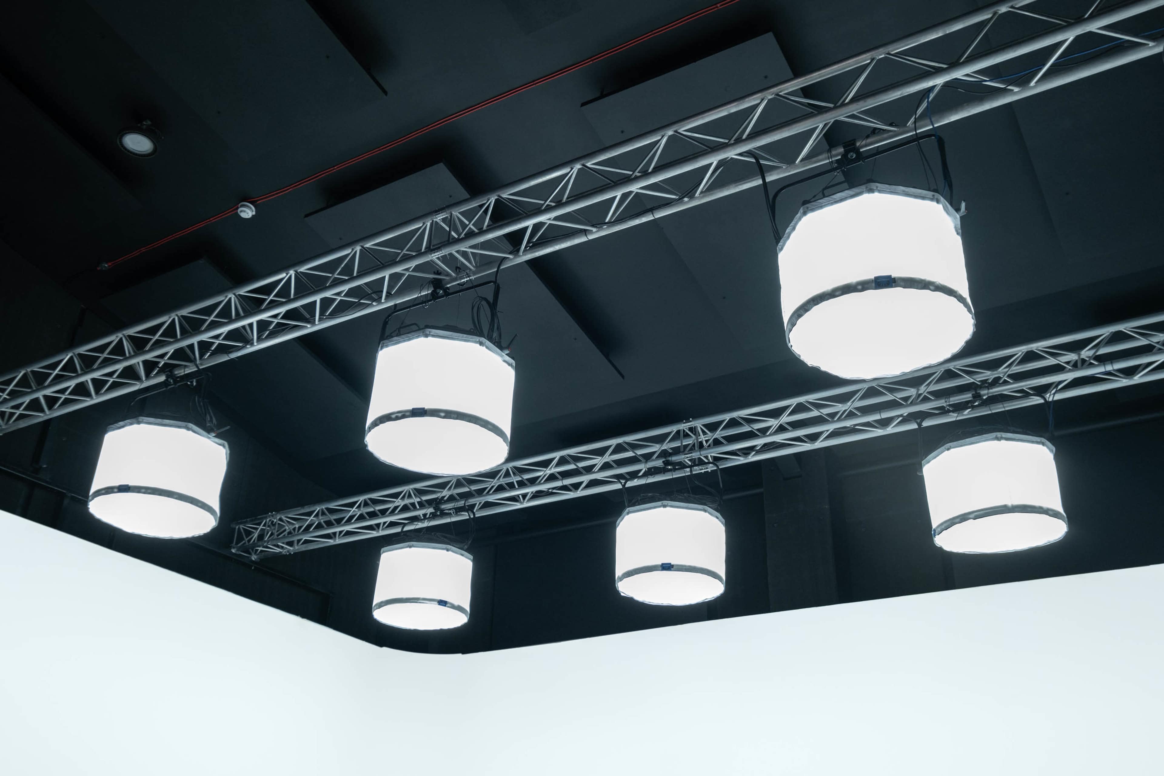 A series of six large, round lights hanging from a metal truss in a studio space.