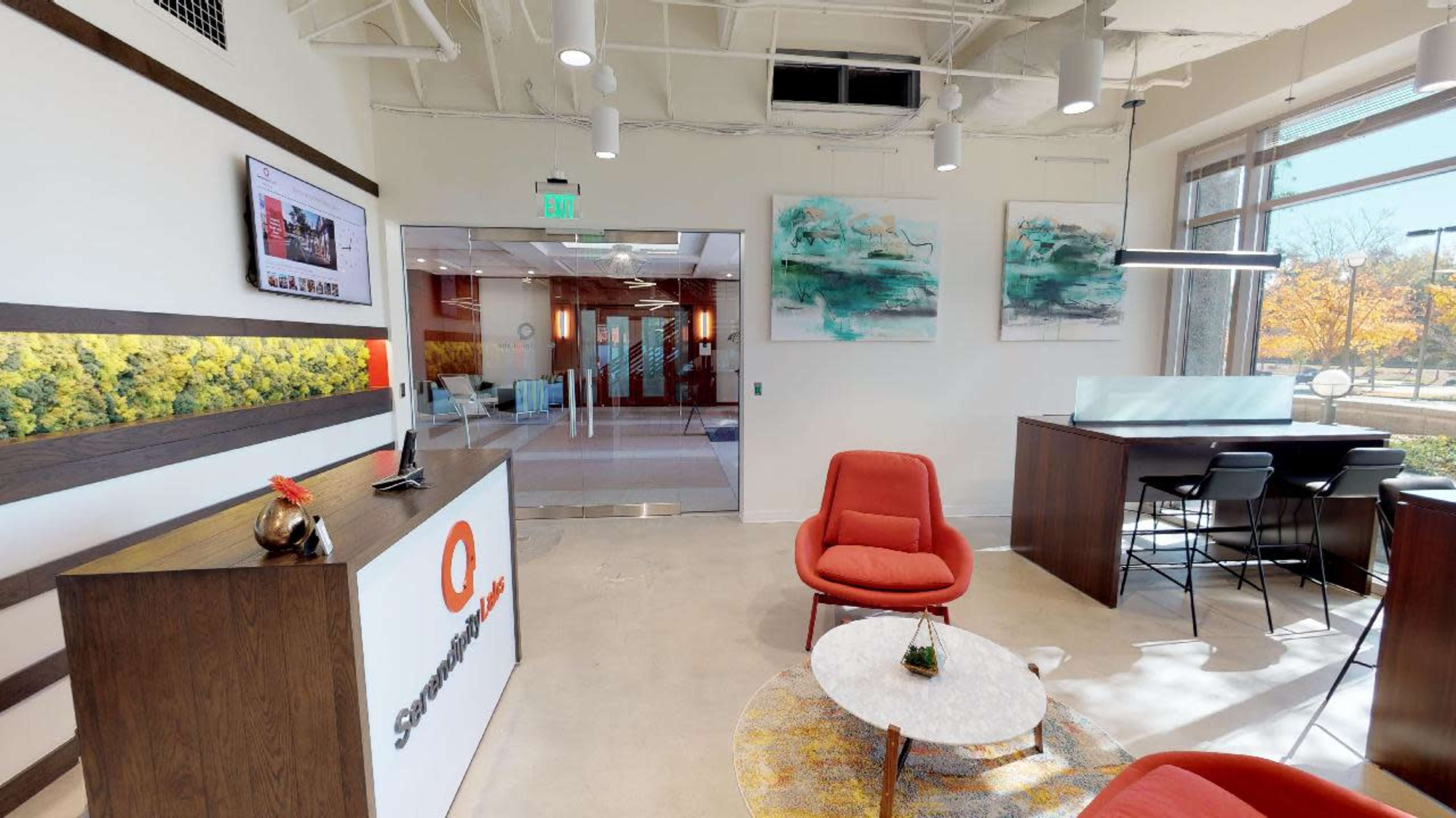 The image shows a modern office reception area featuring a sleek front desk, red chairs, a circular coffee table, and abstract artwork on the walls.