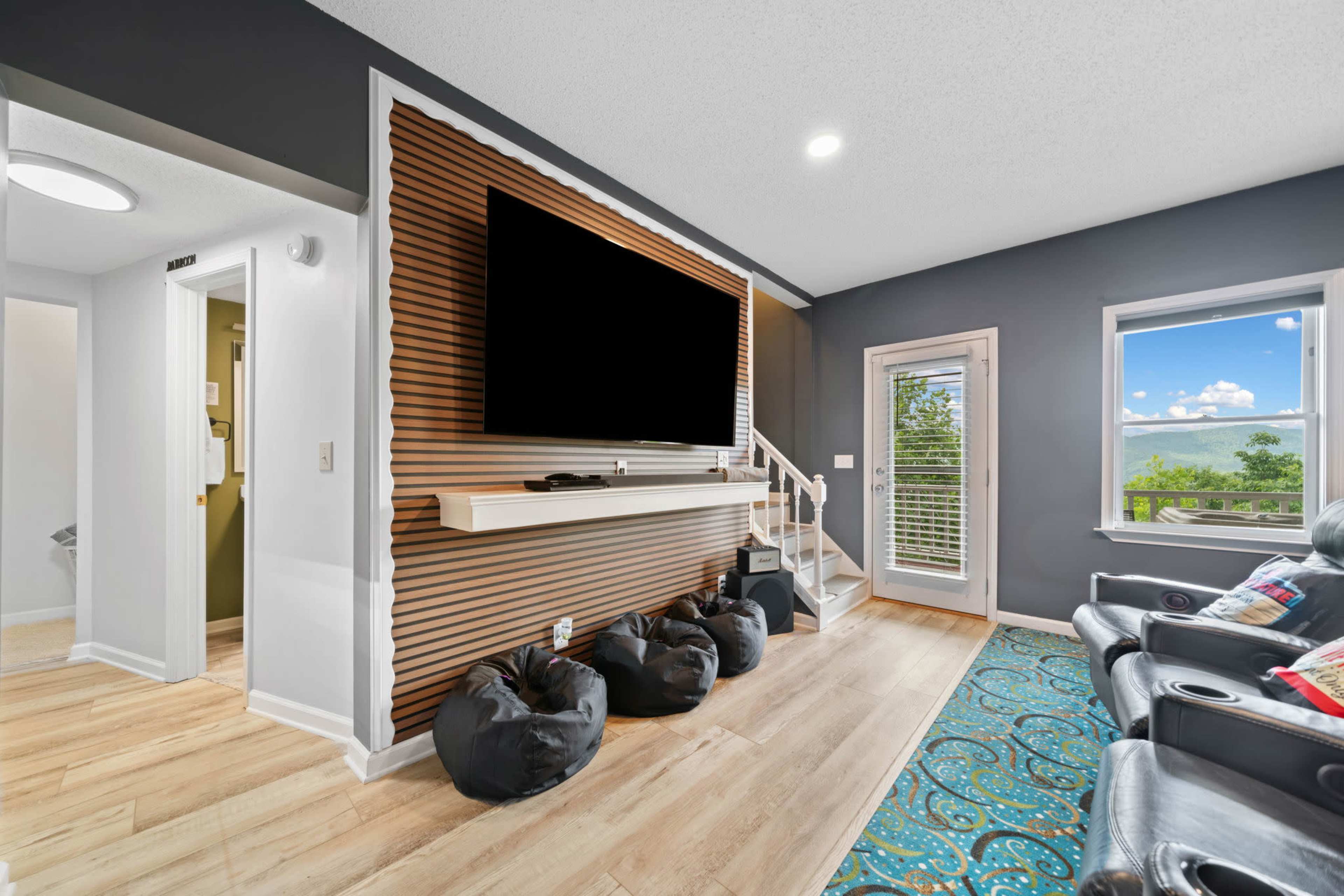 The image shows a modern living room with a large wall-mounted television, a wooden accent wall, and black bean bag chairs arranged on a light wooden floor.