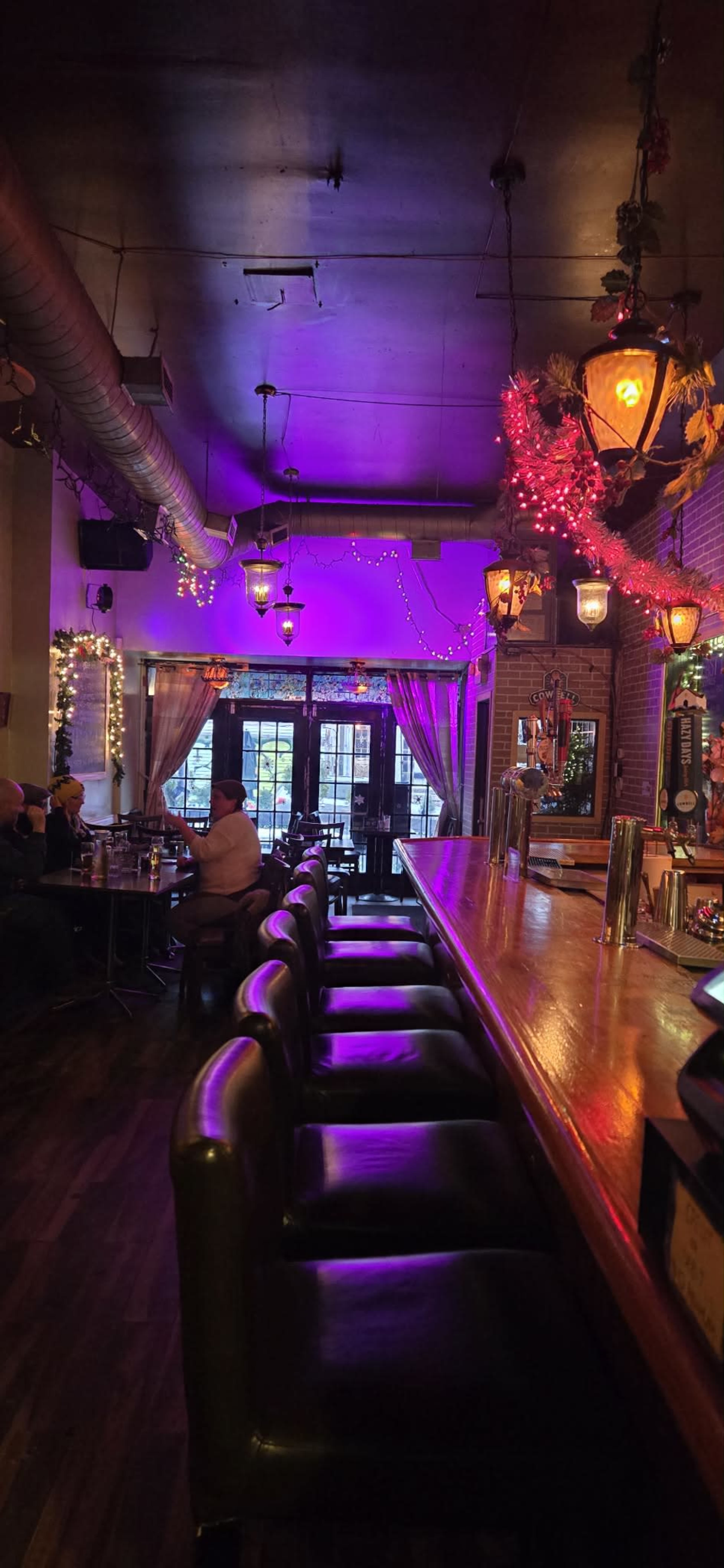 The image shows a dimly lit bar with a long wooden counter, lined with leather stools, and decorative lights overhead.