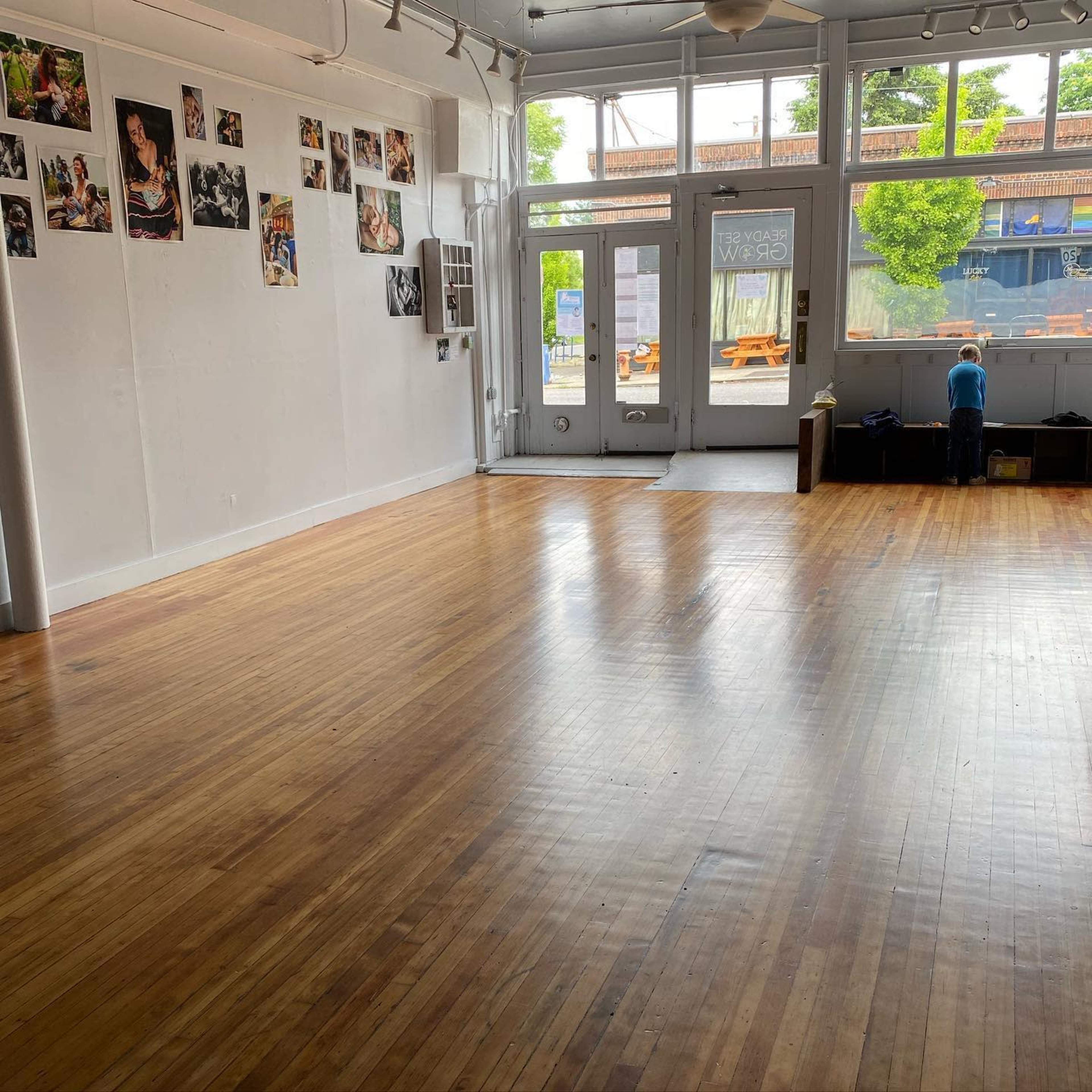A spacious, vacant room with wooden flooring, large windows, and photographs mounted on the walls.
